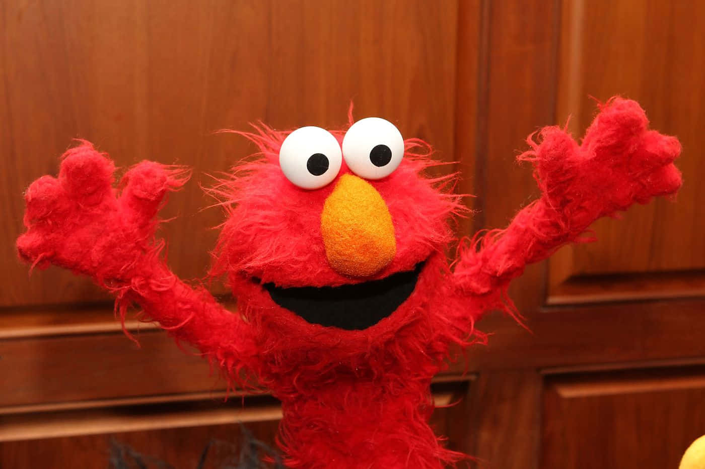 Elmo 桌布 壁紙 — 一隻開心嘅紅色毛絨公仔，有大白色眼睛同橙色鼻，喺木背景前開心咁揮手。