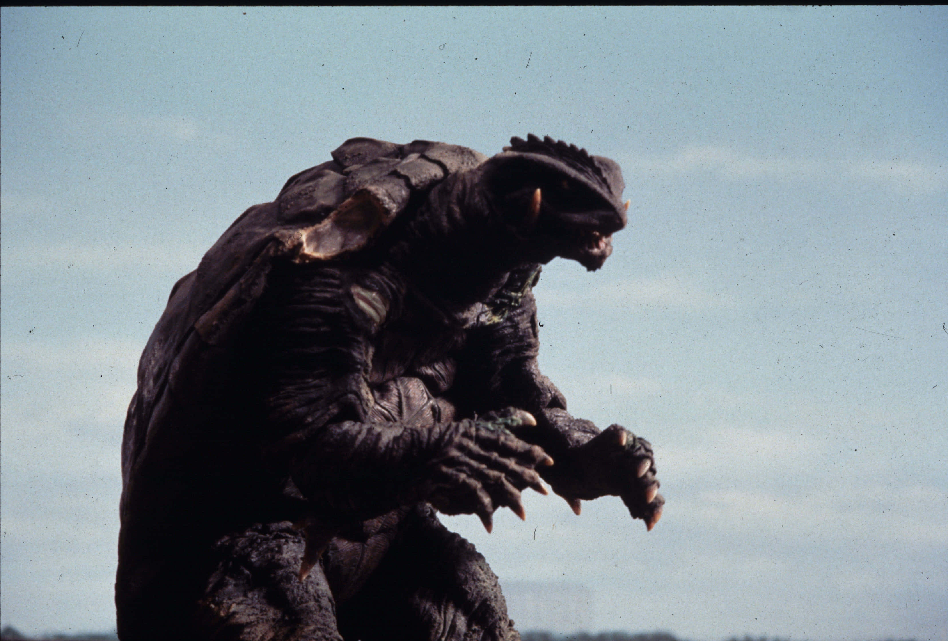 Gamera 桌布 壁紙 — 一隻高聳的深灰色怪獸，擁有裝甲外殼和尖刺頭部，對著蒼白的天空咆哮。