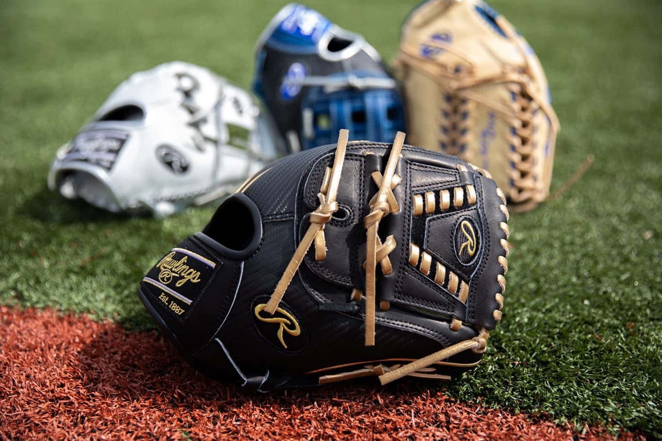 Baseball Gloves 桌布 壁紙 — 一隻黑色Rawlings棒球手套放在紅土上，旁邊有綠色草地，背景還有另外三隻手套。