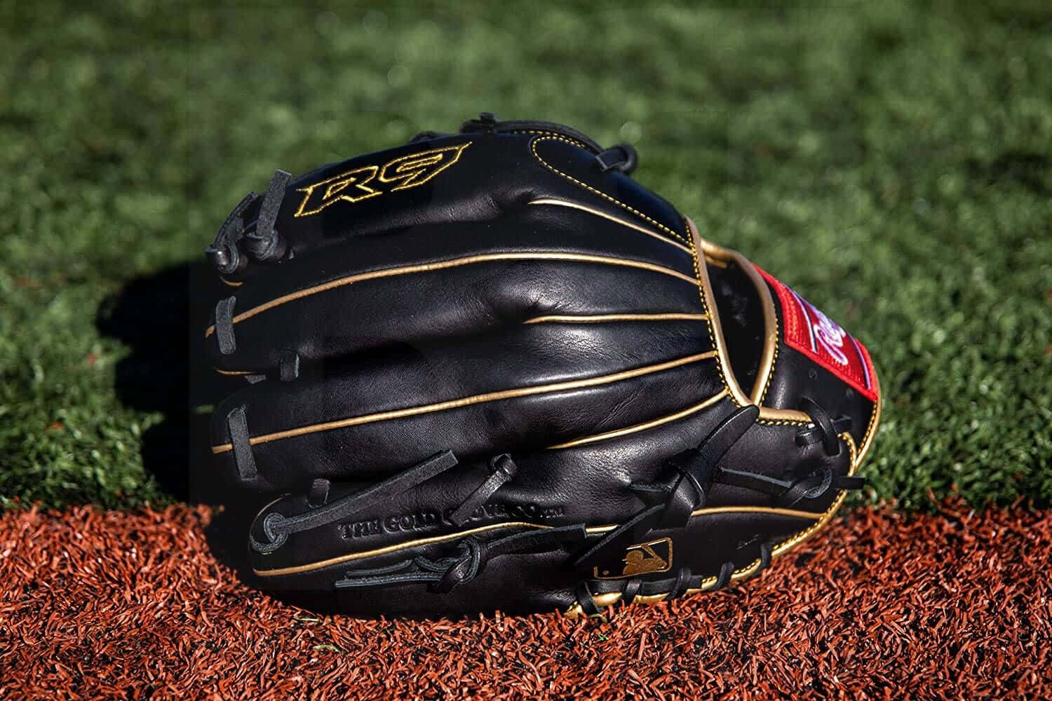 Baseball Gloves 桌布 壁紙 — 一隻黑色棒球手套帶金色縫線，放在紅色泥土上，背景是鮮綠色草地，在明亮陽光下。
