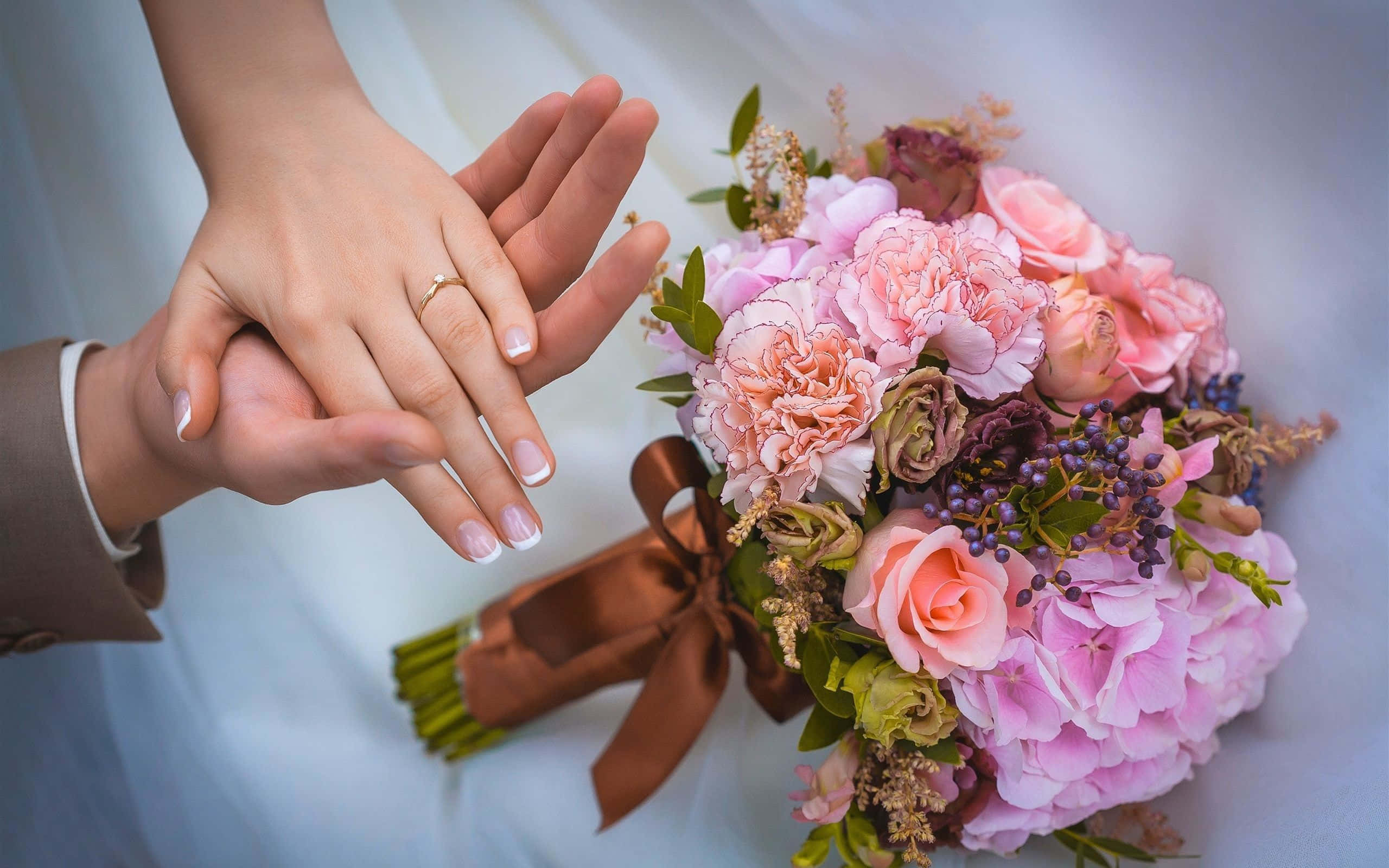 Wedding Flowers 桌布 壁紙 — 一雙交織的手的特寫，其中一隻手戴著結婚戒指，旁邊是一個用棕色絲帶裝飾的茂盛粉紅色花束。
