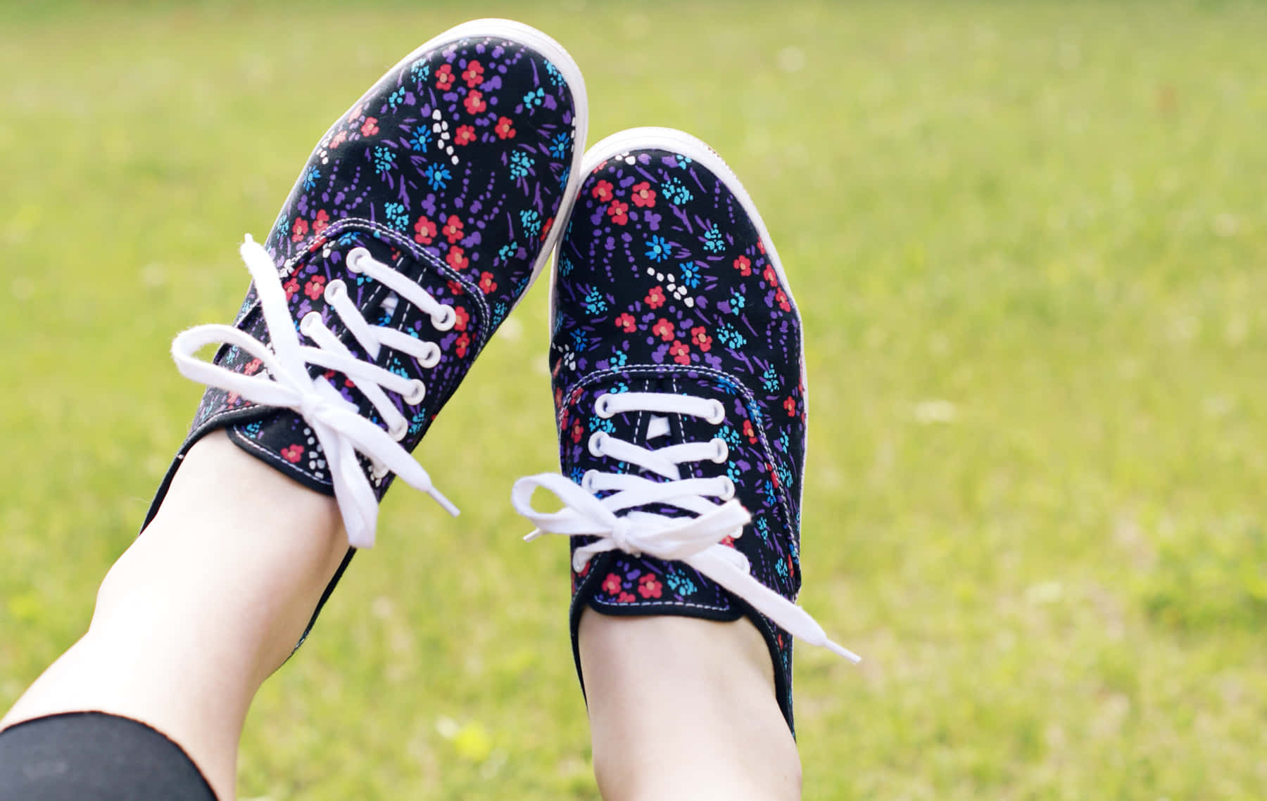 Cute Shoes 桌布 壁紙 — 一雙穿著花卉圖案運動鞋的腳，在明亮的日光下隨意地放在充滿活力的綠色草地上。