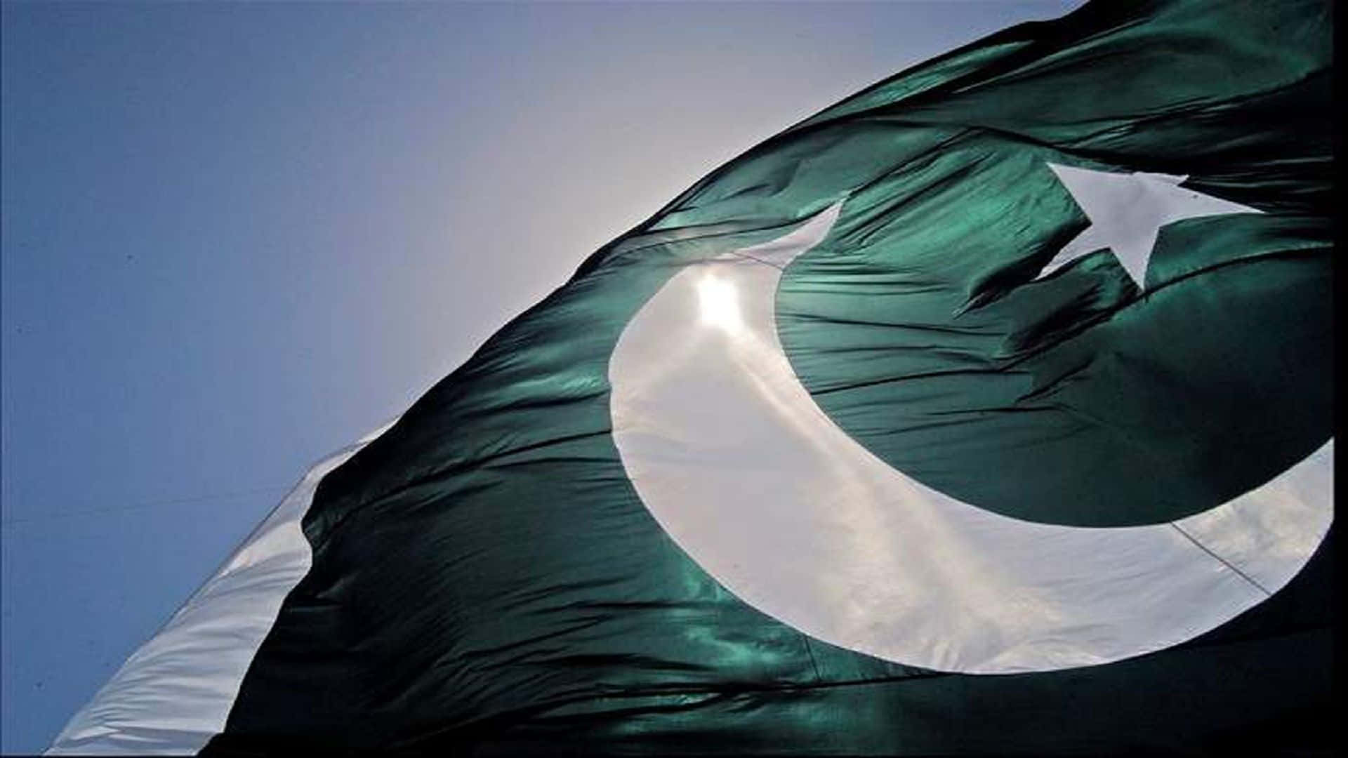 Pakistan 桌布 壁紙 — 一面大型巴基斯坦國旗在清澈的藍天下動態飄揚，陽光在布料上閃爍。