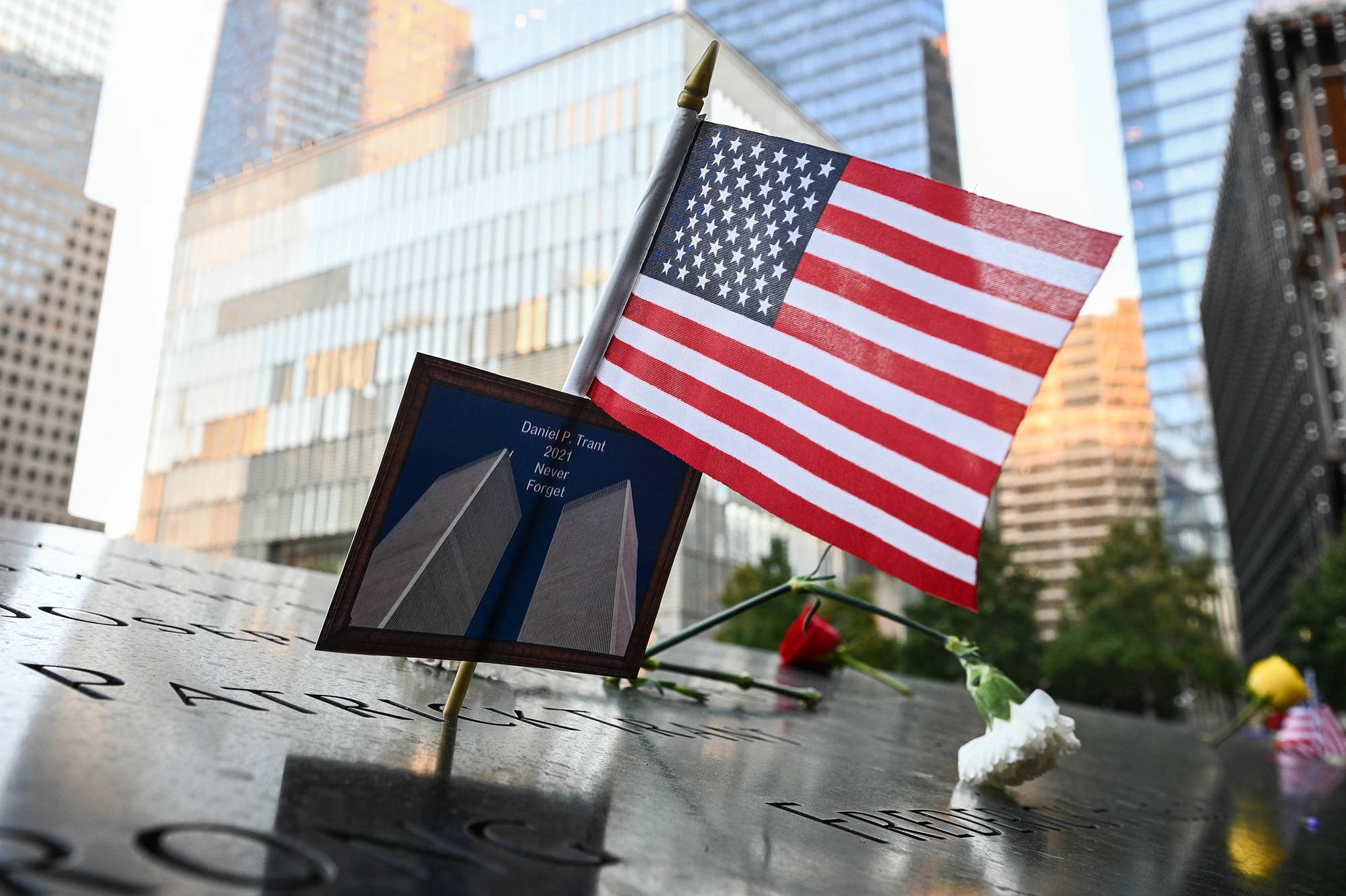 911 Memorial 桌布 壁紙 — 一面小型美國國旗和一幅雙子塔的裱框照片放在反射的911紀念池上，四周環繞著摩天大樓。