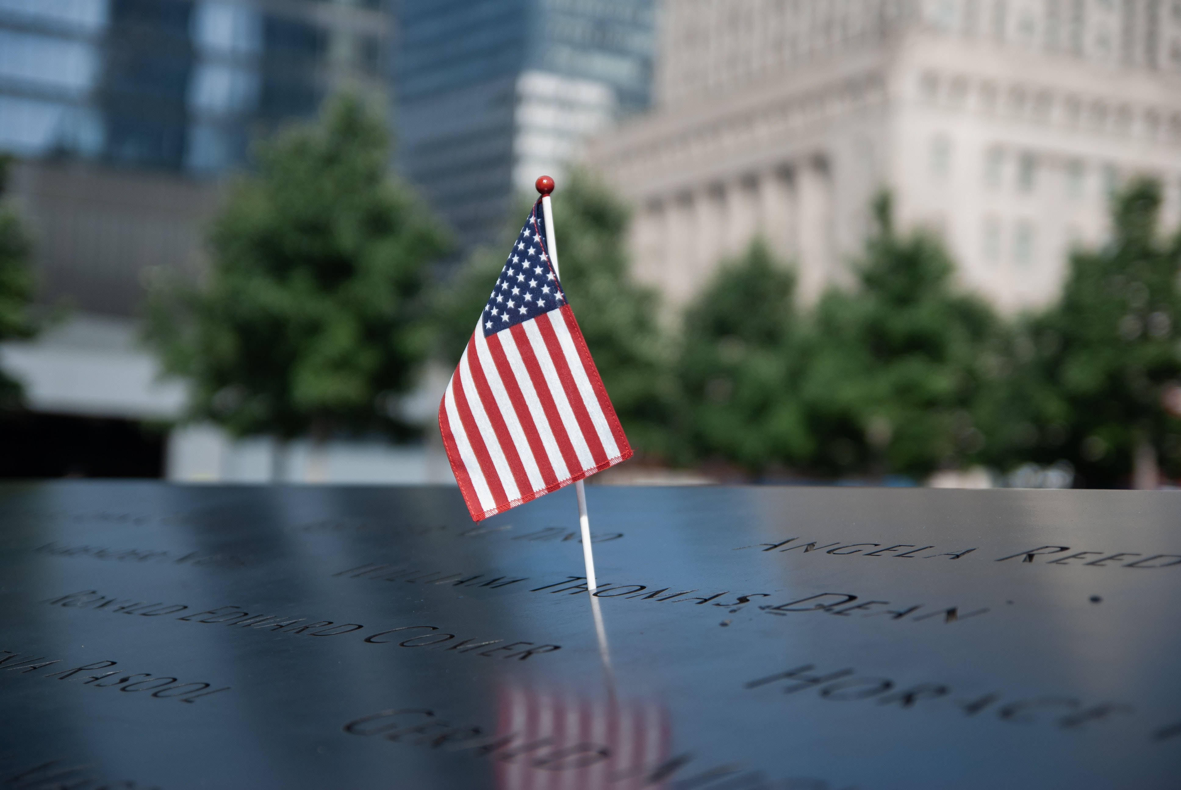 911 Memorial 桌布 壁紙 — 一面小型美國國旗自豪地矗立在刻有名字的反光紀念表面上，背景是模糊的城市建築和綠樹。