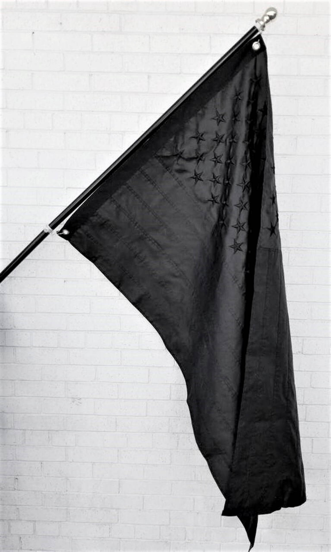 Black American Flag 桌布 壁紙 — 一面黑色旗幟上縫有星星，無力地掛在旗桿上，背景是白色磚牆。