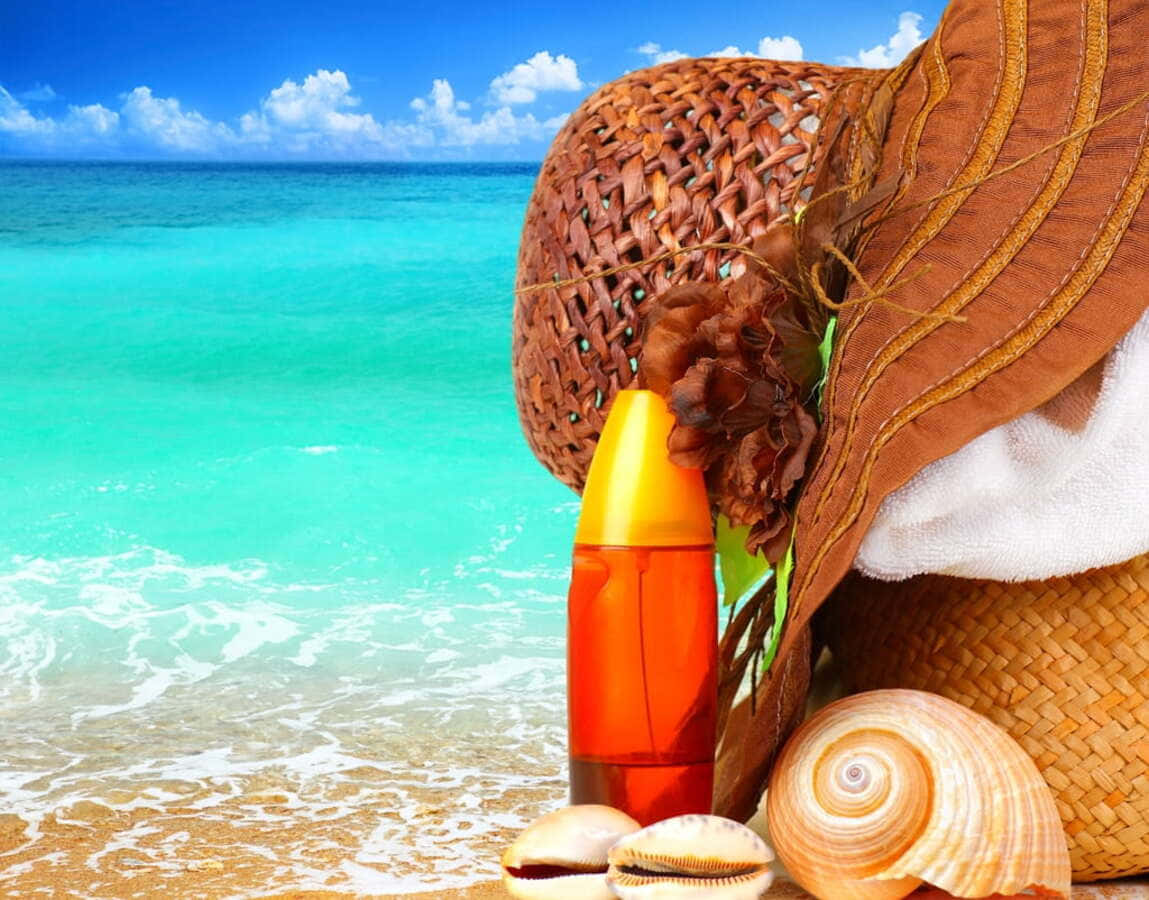 Beach Bag 桌布 壁紙 — 一頂太陽帽、防曬霜瓶子和貝殼放在金色沙灘上，旁邊是碧綠的海浪，上方是明亮的藍天。