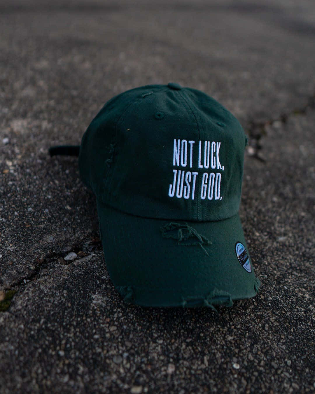 Hat 桌布 壁紙 — 一頂深綠色破舊的棒球帽，上面有白色文字寫著 not luck just god，放在破裂的瀝青路面上。