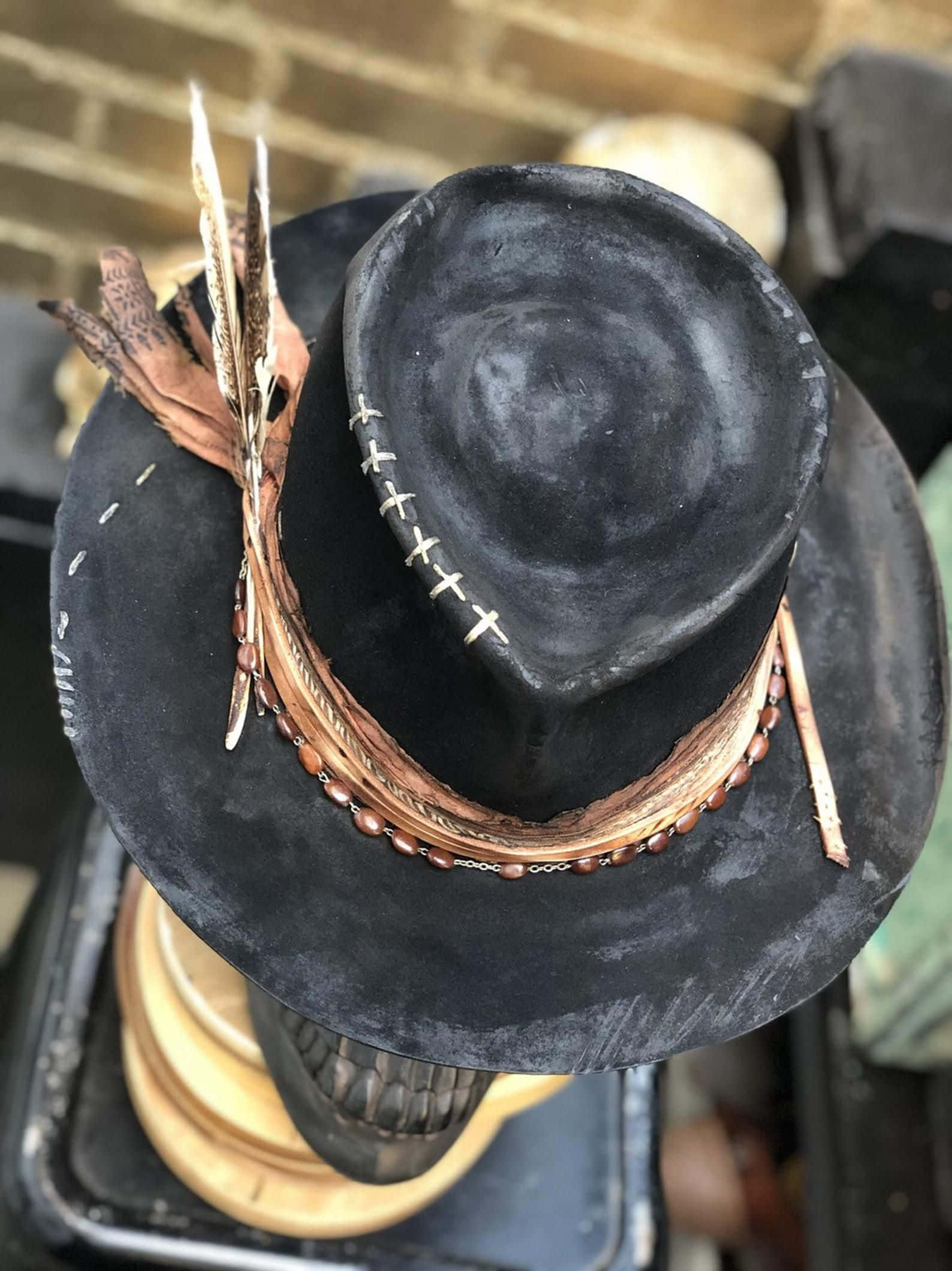 Hat 桌布 壁紙 — 一頂破舊的黑色牛仔帽，飾有羽毛、珠子和皮帶，放在一個骷髏形狀的支架上，背景是磚牆。
