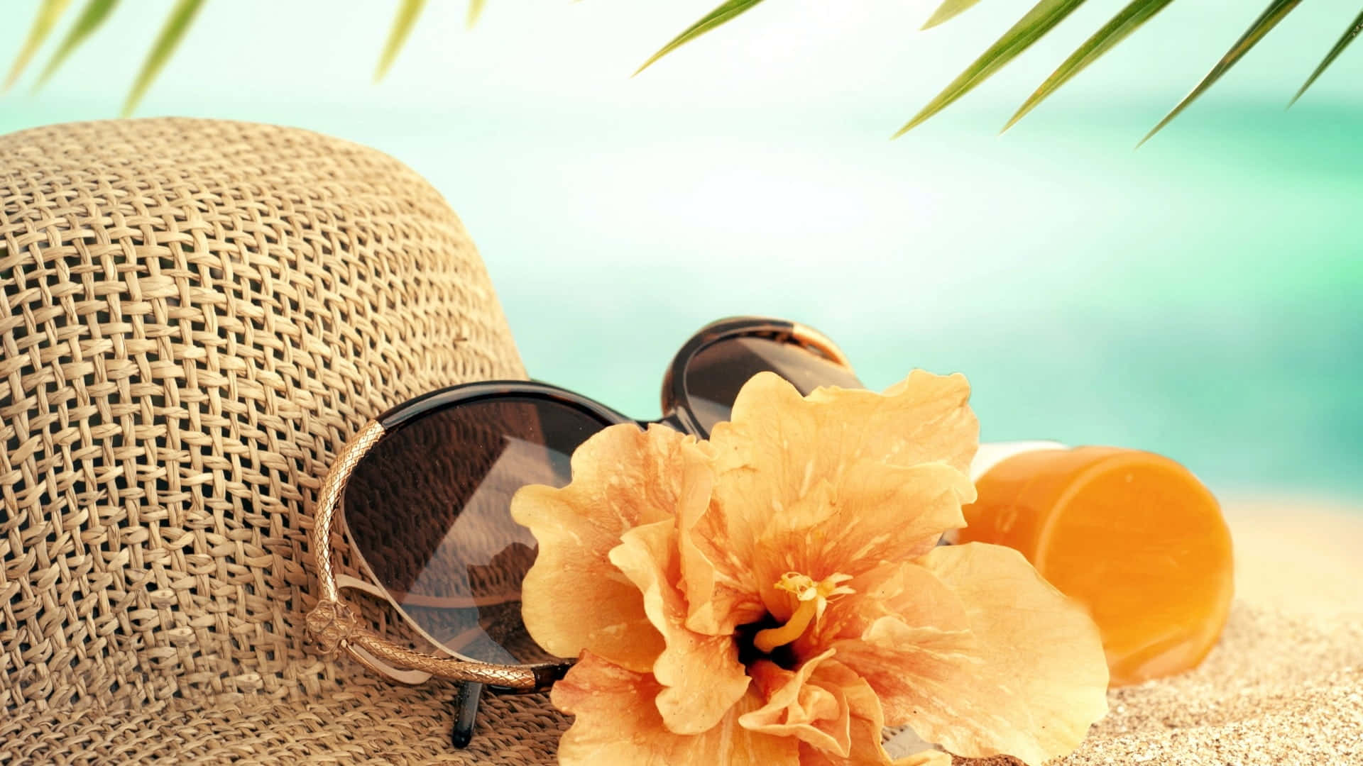 Beach Accessories 桌布 壁紙 — 一頂草帽、太陽眼鏡和橙色木芙蓉花放在沙灘上，棕櫚葉下，背景是碧綠的海洋。