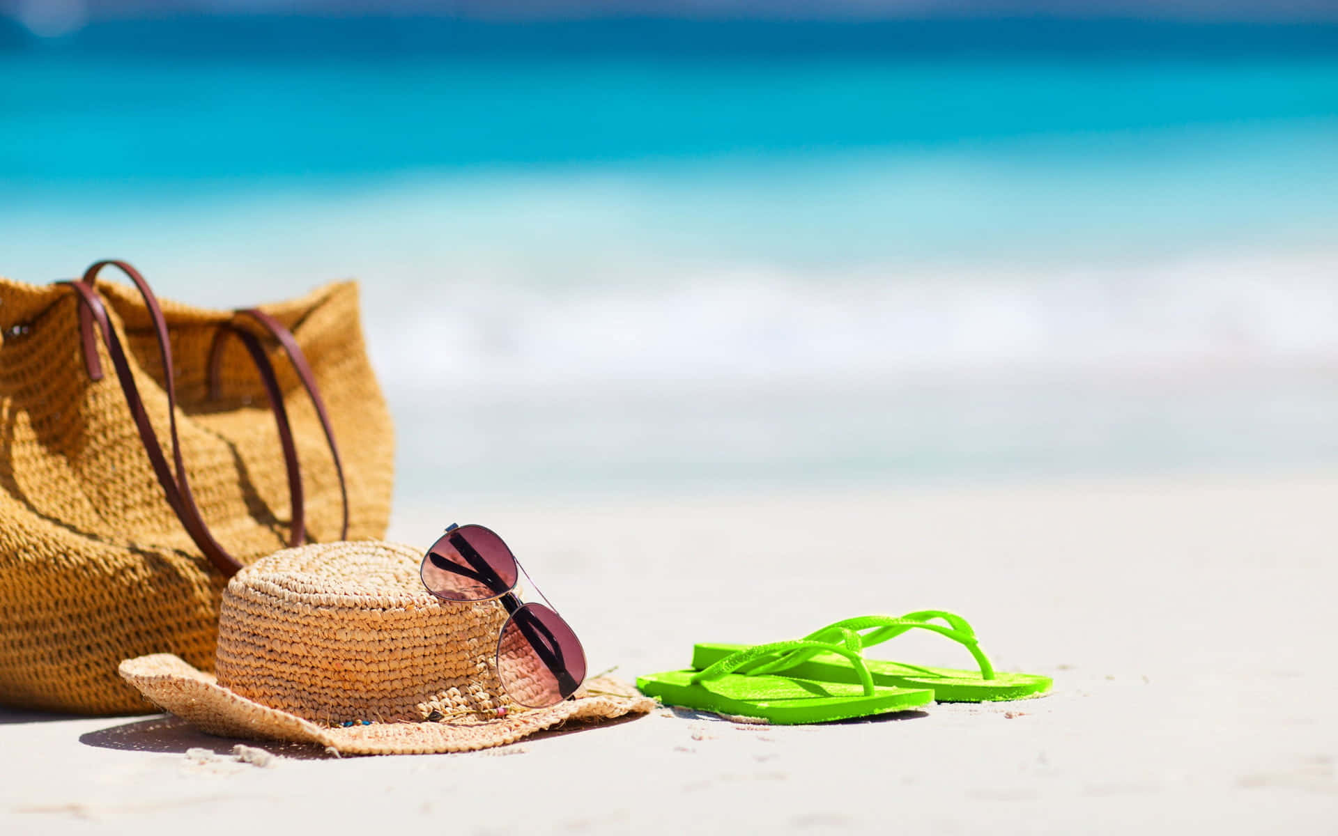 Beach Hat 桌布 壁紙 — 一頂草帽、太陽眼鏡和鮮綠色人字拖放在白色沙灘上，旁邊有一個編織沙灘袋，背景是碧綠色的海浪。