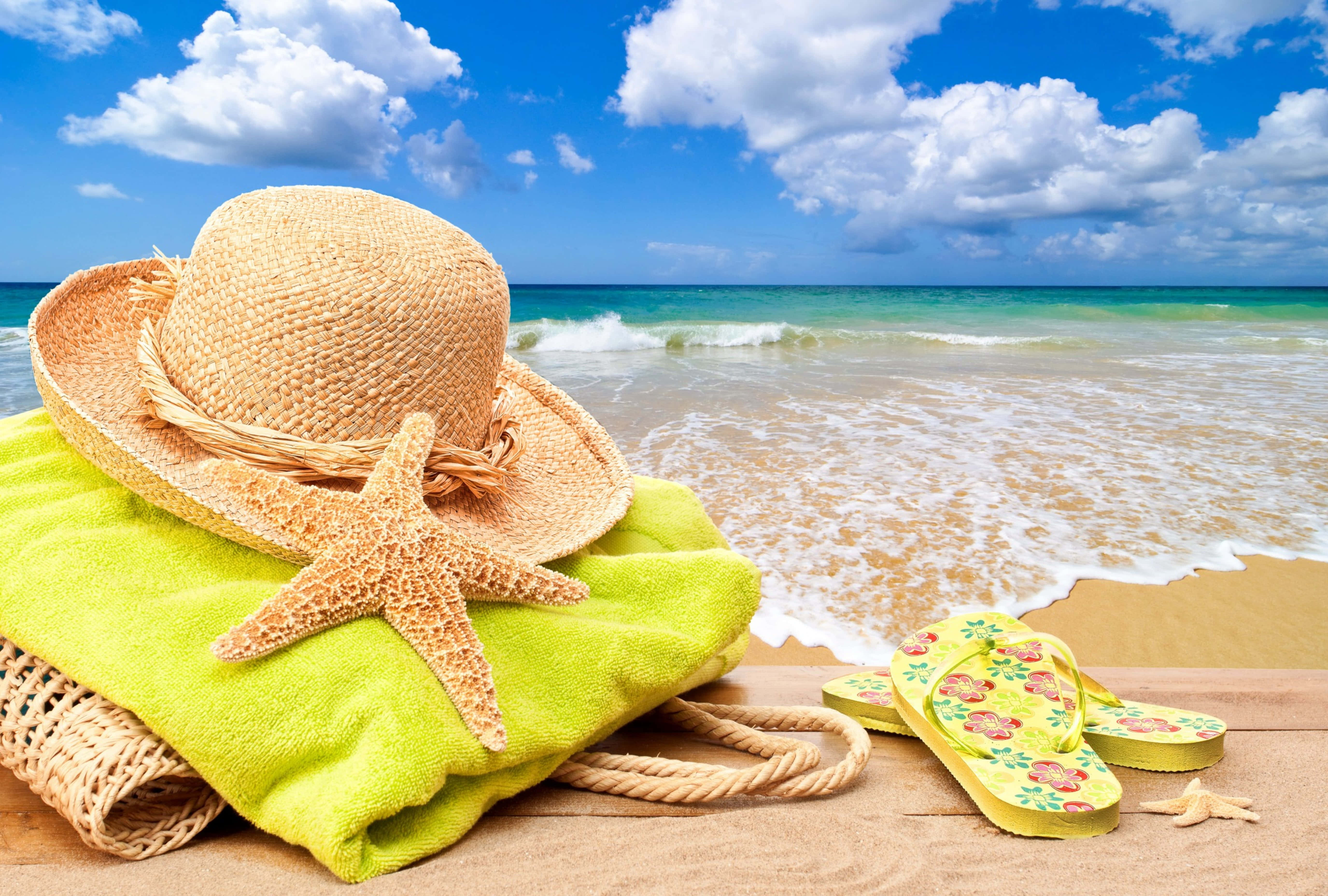 Beach Accessories 桌布 壁紙 — 一頂草帽、海星和花卉人字拖放在亮綠色毛巾上，旁邊是海浪，天空蔚藍，白雲朵朵。
