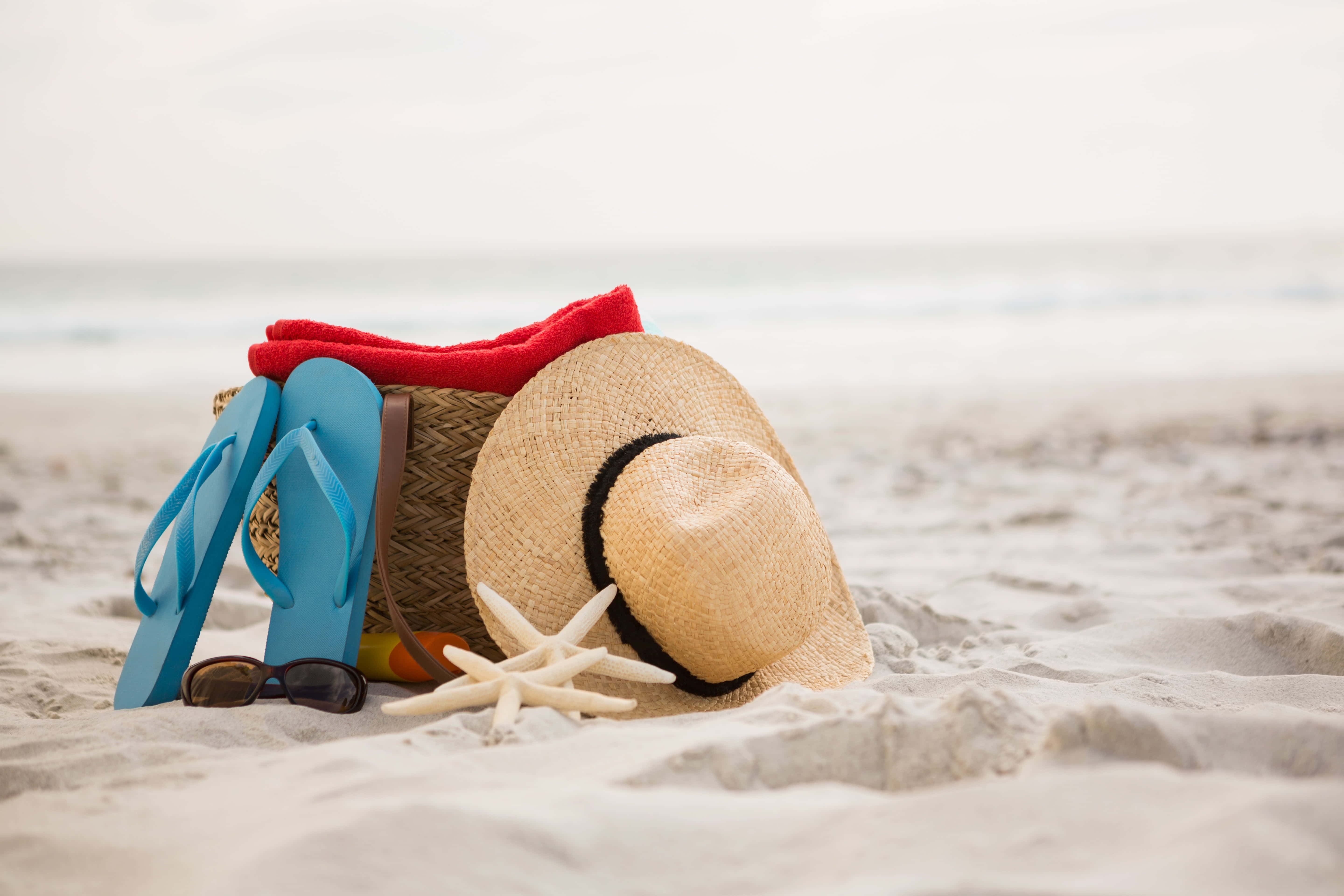 Beach Bag 桌布 壁紙 — 一頂草帽、藍色人字拖和太陽眼鏡放在沙灘上，旁邊有海星和紅色毛巾，靠近海洋。