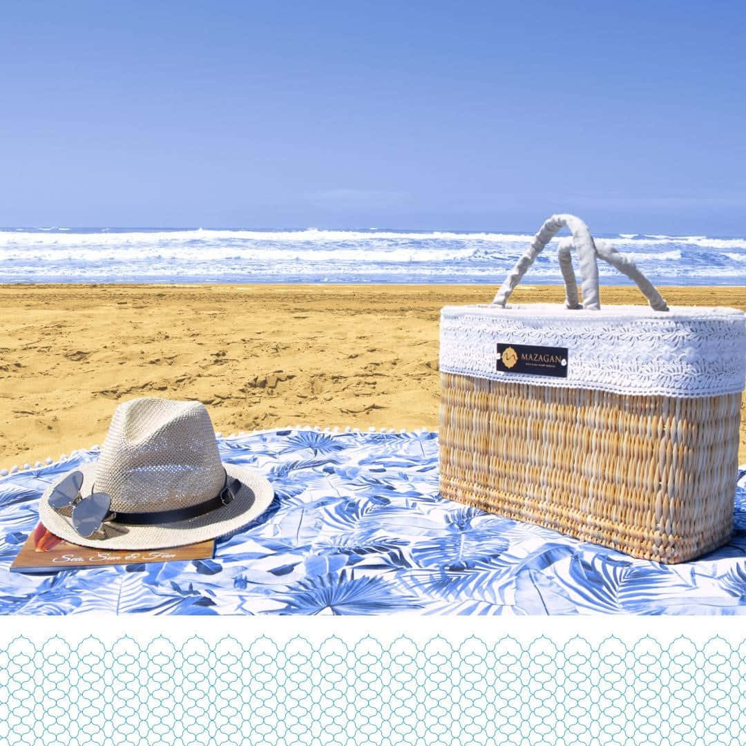 Beach Picnic 桌布 壁紙 — 一頂草帽和一個編織籃子放在一條藍色棕櫚圖案沙灘毛巾上，旁邊是清澈藍天下拍岸的海浪。