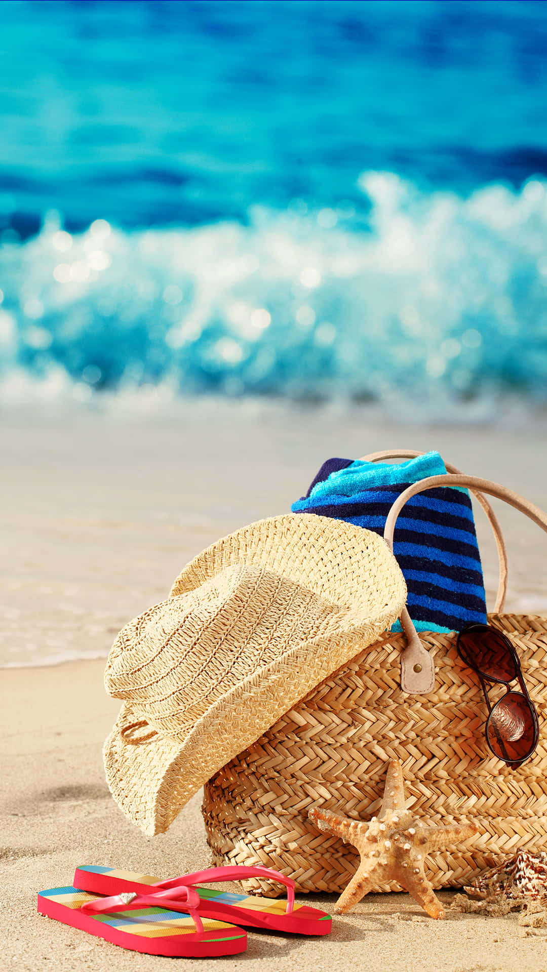 Beach Accessories 桌布 壁紙 — 一頂草帽和色彩繽紛的拖鞋放在一個編織沙灘袋旁邊，袋上有太陽眼鏡和海星，擱在金色沙灘上，附近有碧綠色的海浪拍打。