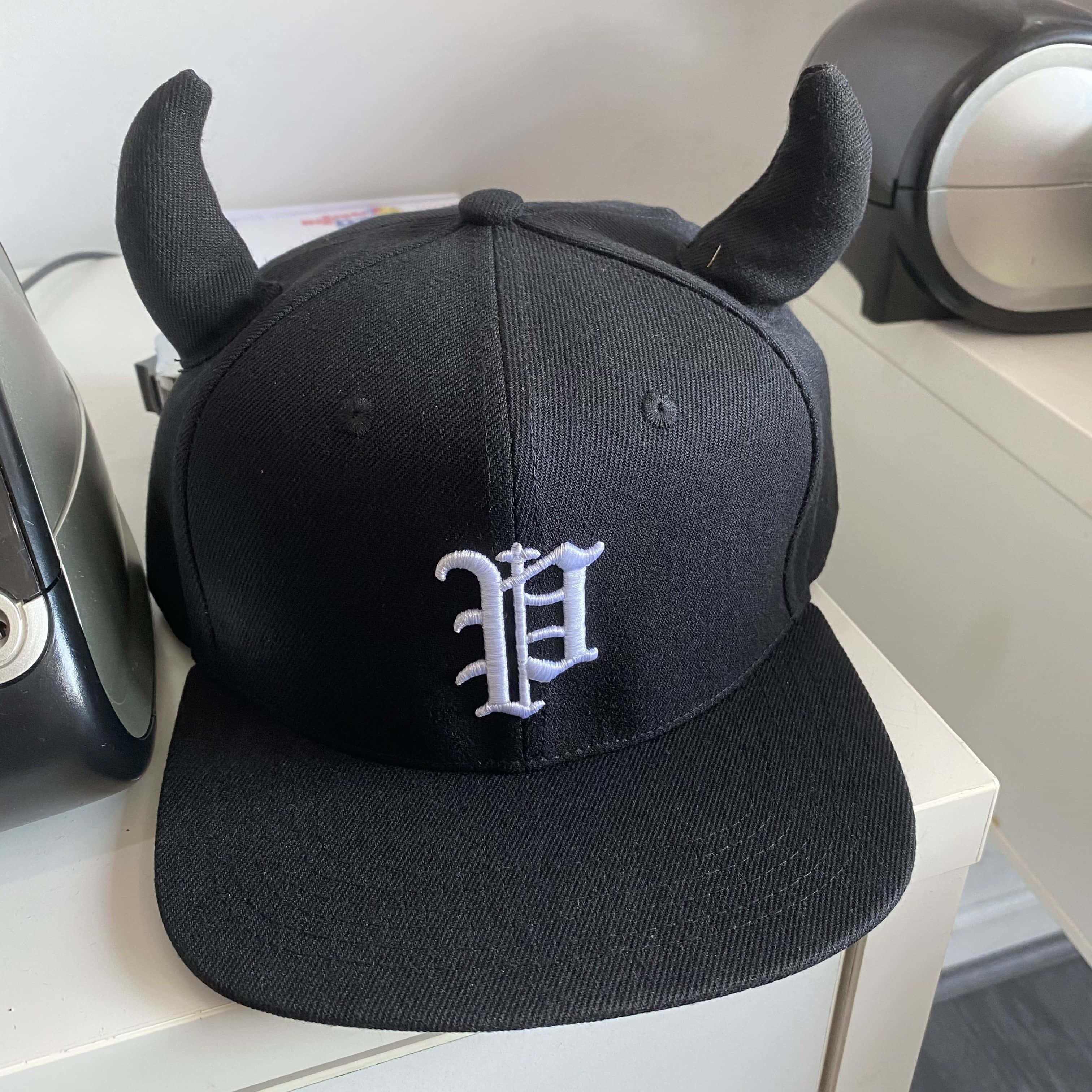 Hat 桌布 壁紙 — 一頂黑色 snapback 帽，帶有魔鬼角和白色刺繡字母 D，放在白色架子上，旁邊有電器。
