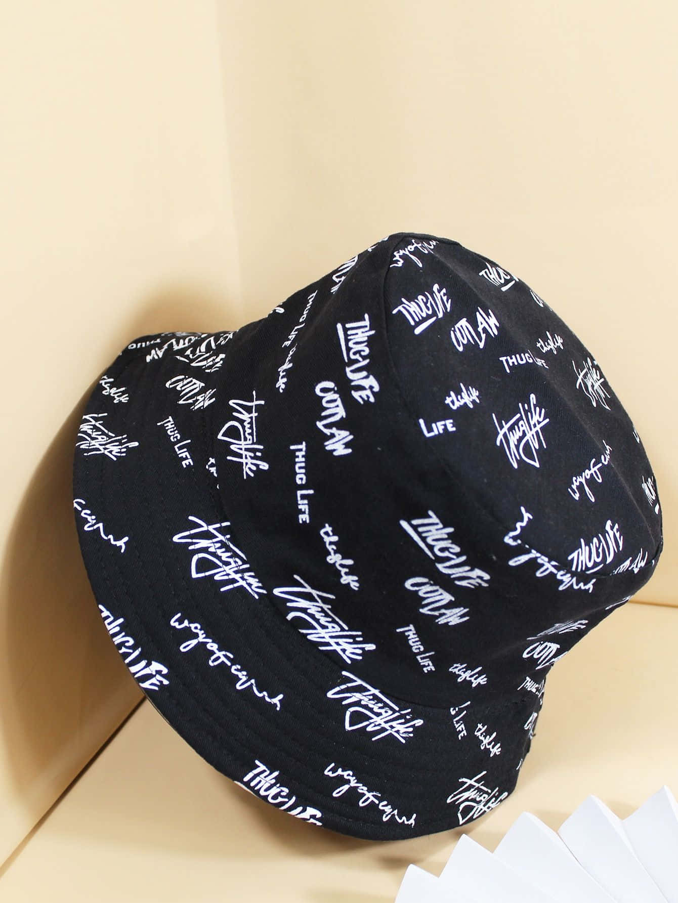 Hat 桌布 壁紙 — 一頂黑色漁夫帽上佈滿白色草書文字，靠在一面米色牆上，附近有白色摺紙。