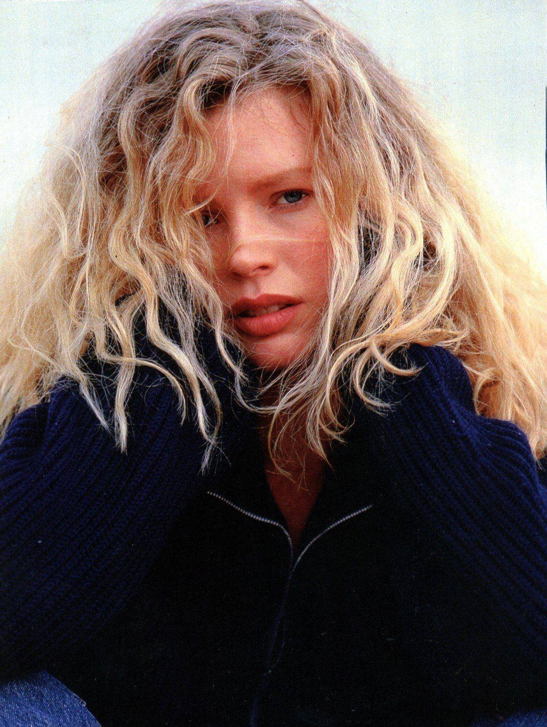 Kim Basinger In A Simple Sweater 壁紙 — 一頭凌亂金髮的女人，穿著深藍色羅紋毛衣，在柔和模糊的背景前凝視著。