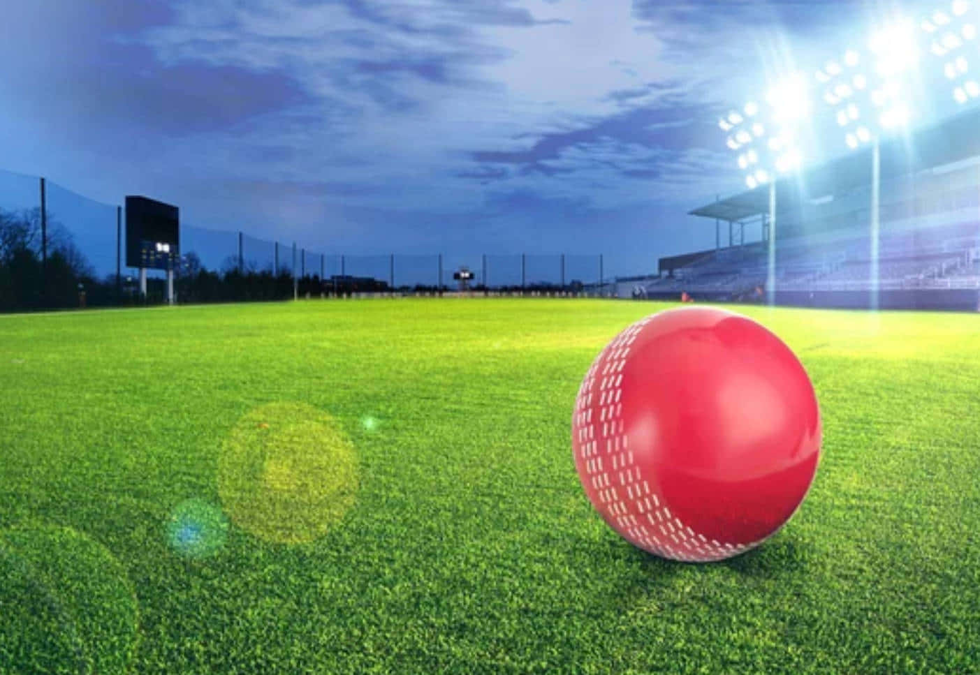 Cricket Ground 桌布 壁紙 — 一顆亮面紅色板球在暮色中躺在體育場燈光下的翠綠草地上。