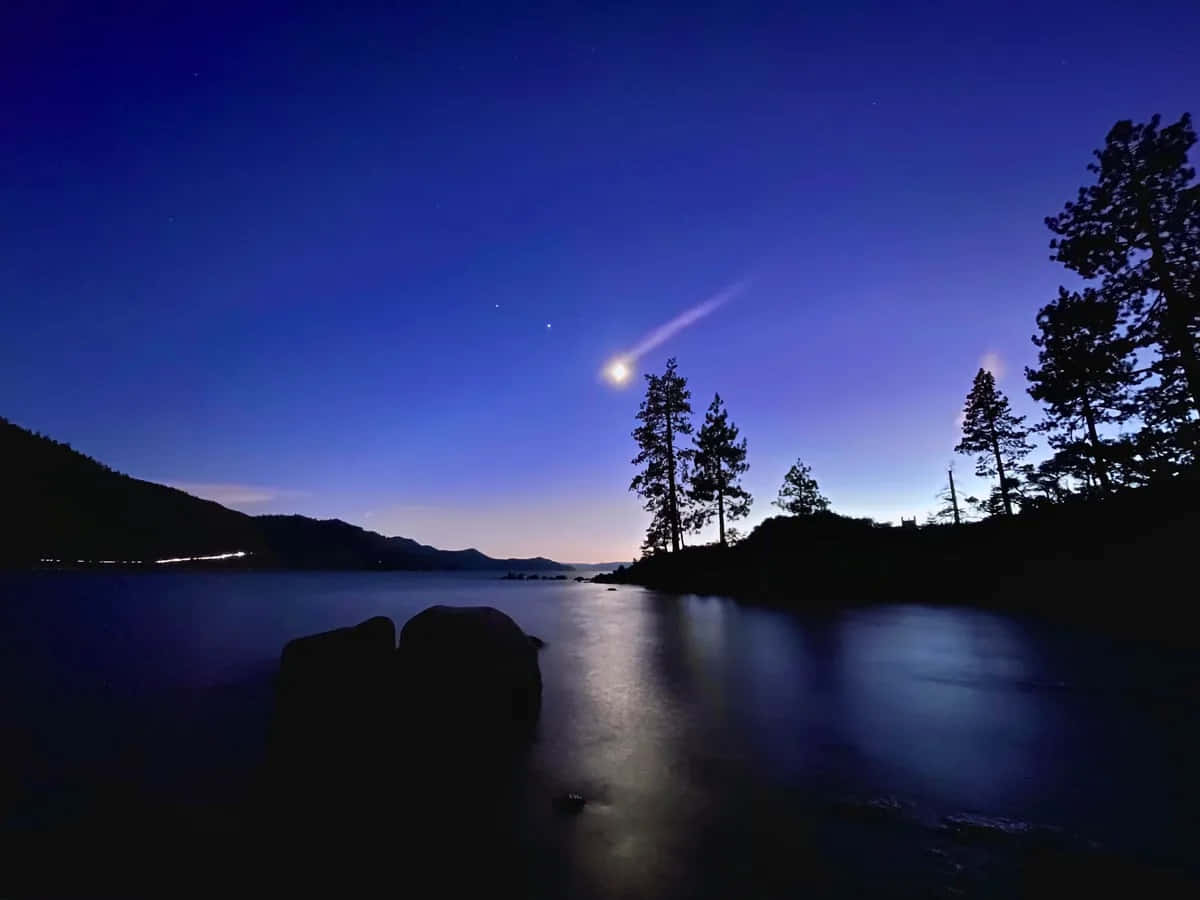 Night Mode 桌布 壁紙 — 一顆彗星劃過繁星點點的暮色天空，下方是平靜的湖泊，四周環繞著剪影般的樹木和遠山。