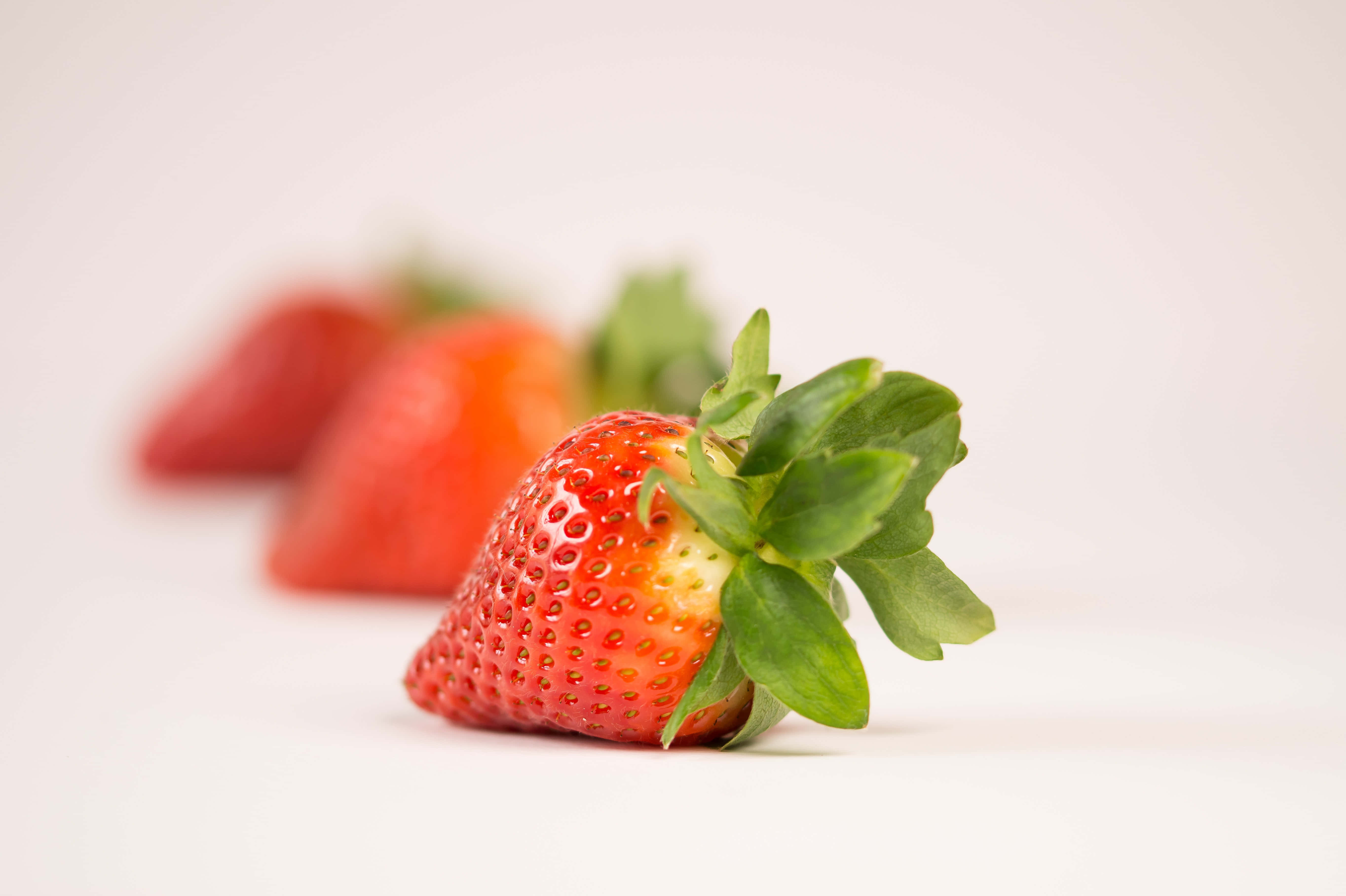 Summer Fruits 桌布 壁紙 — 一顆成熟的草莓，帶有綠葉，在柔和的白色背景上清晰對焦，後面有兩顆模糊的草莓。