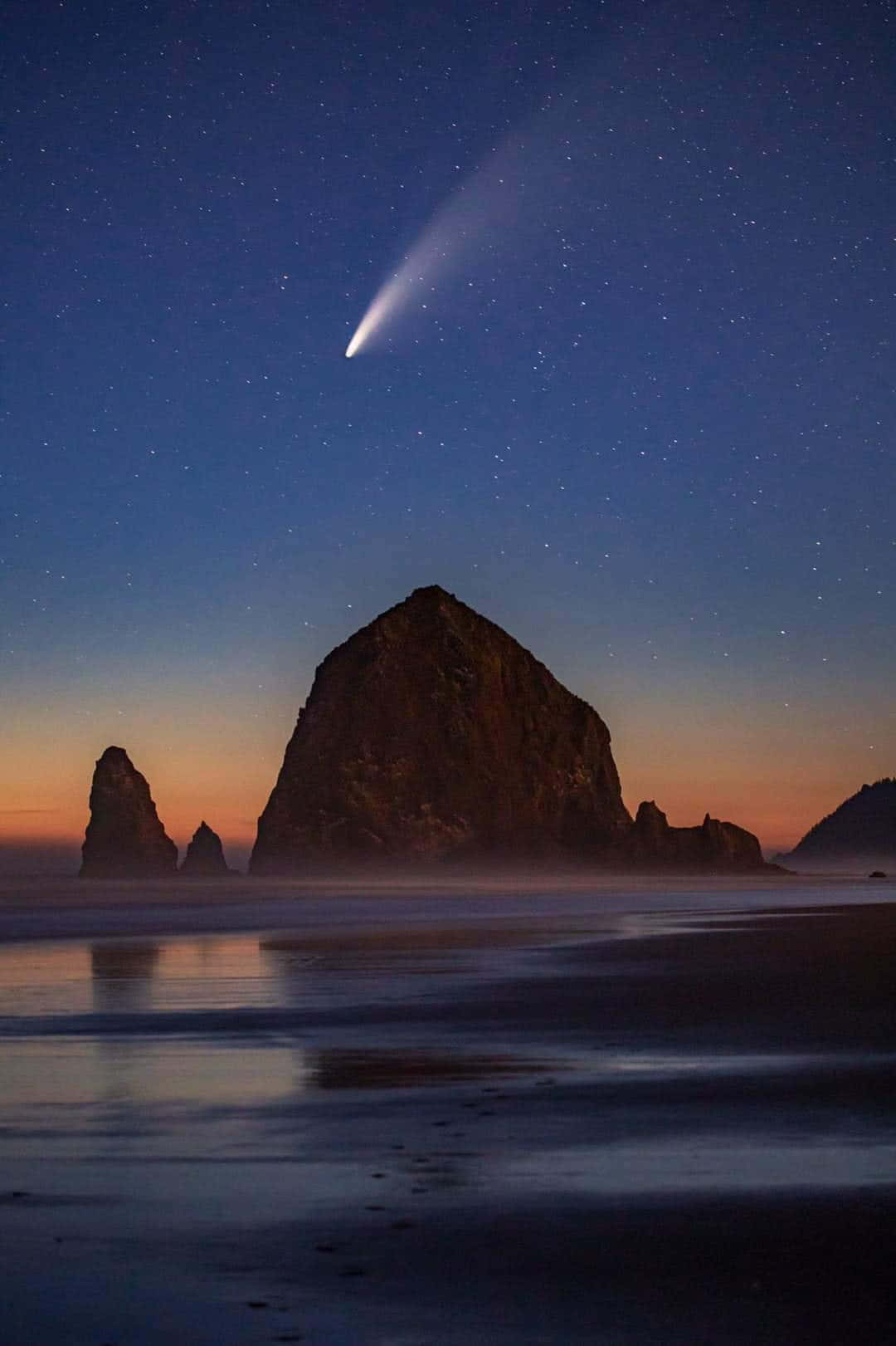 comet 桌布 壁紙 — 一顆明亮的彗星劃過佈滿星星的暮色天空，下方是霧氣籠罩的海岸岩石和寧靜如鏡的水面。