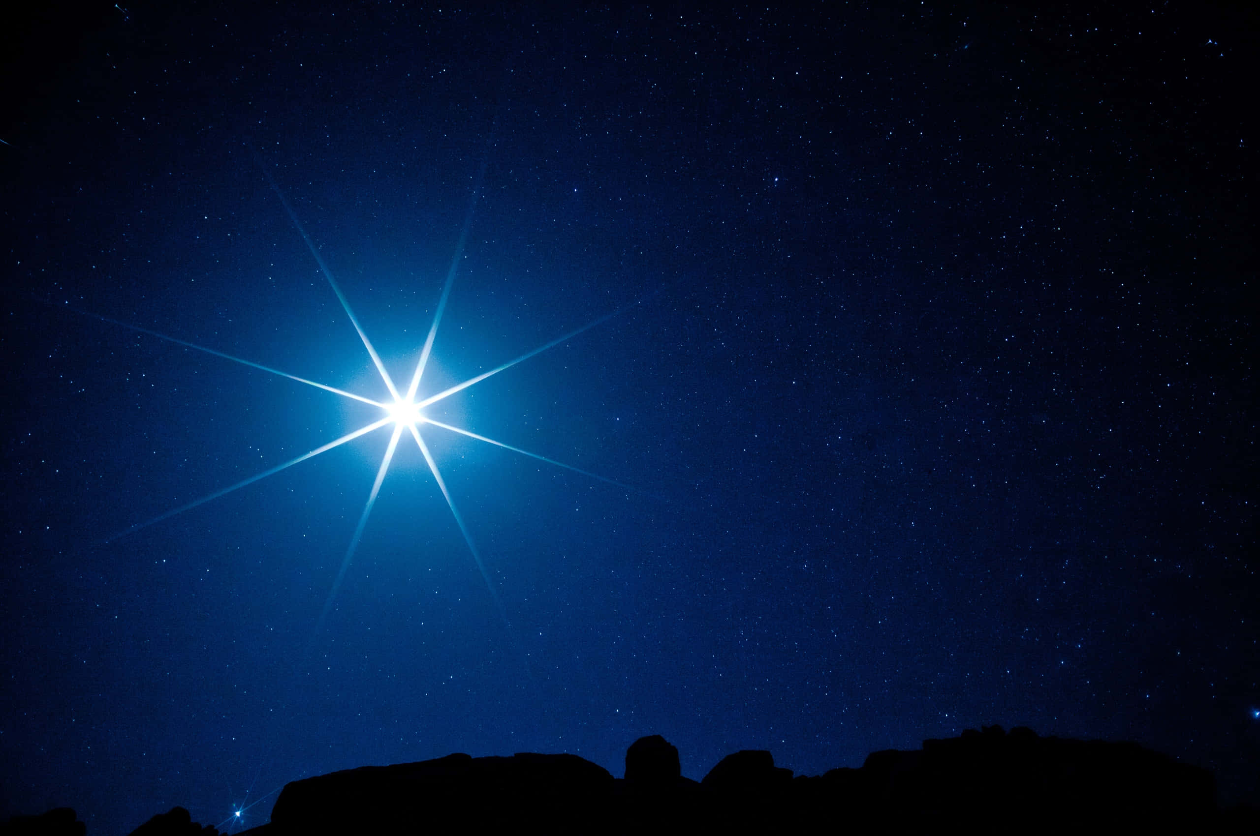 3d Star 桌布 壁紙 — 一顆明亮的星星帶著輻射光束，穿透深藍色的夜空，夜空點綴著星星，下方是黑暗的岩石剪影。