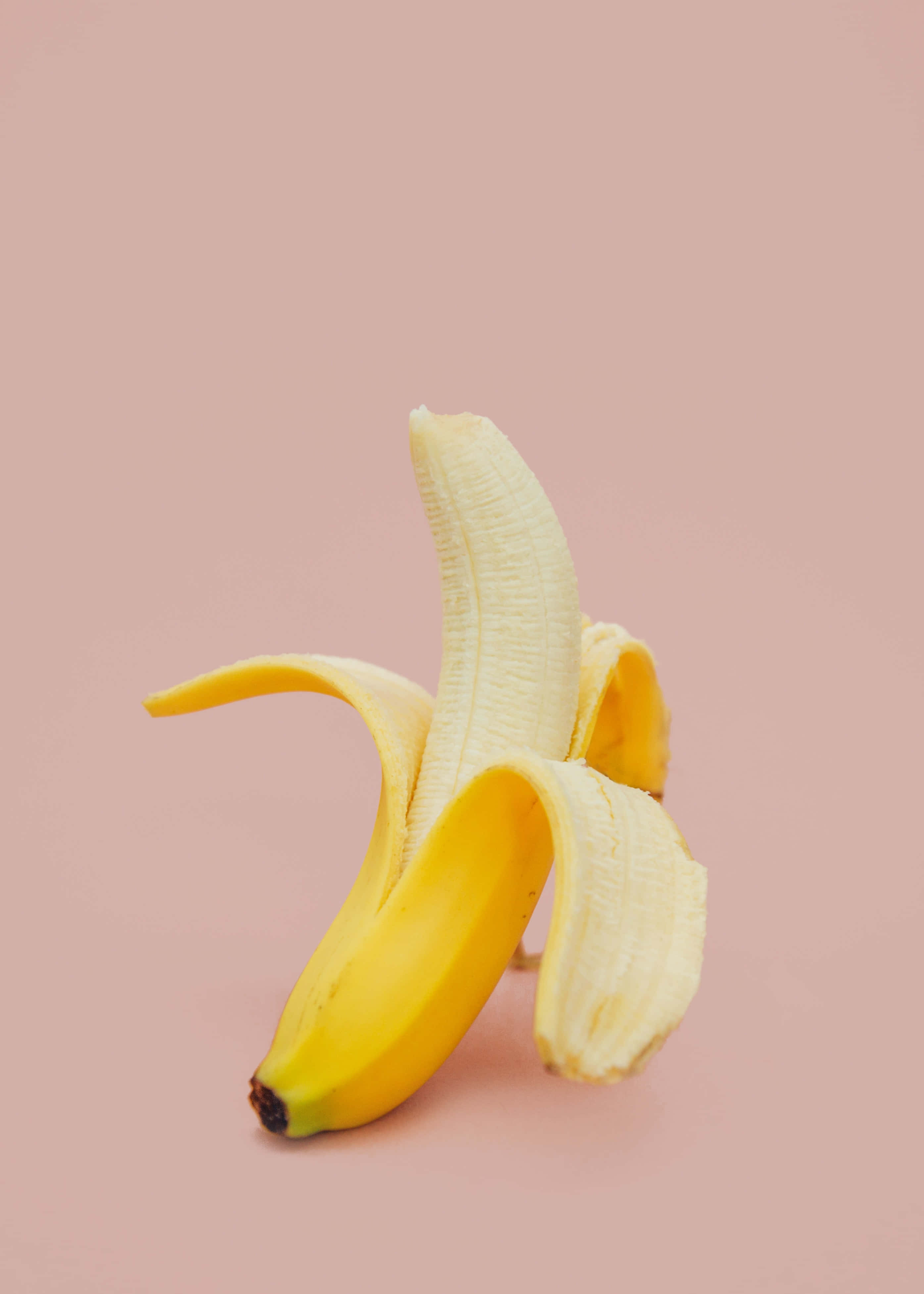 一顆美味的黃色香蕉放在白色盤子上 壁紙 — 單條剝皮香蕉，黃色香蕉皮展開，襯以柔和粉紅色背景。
