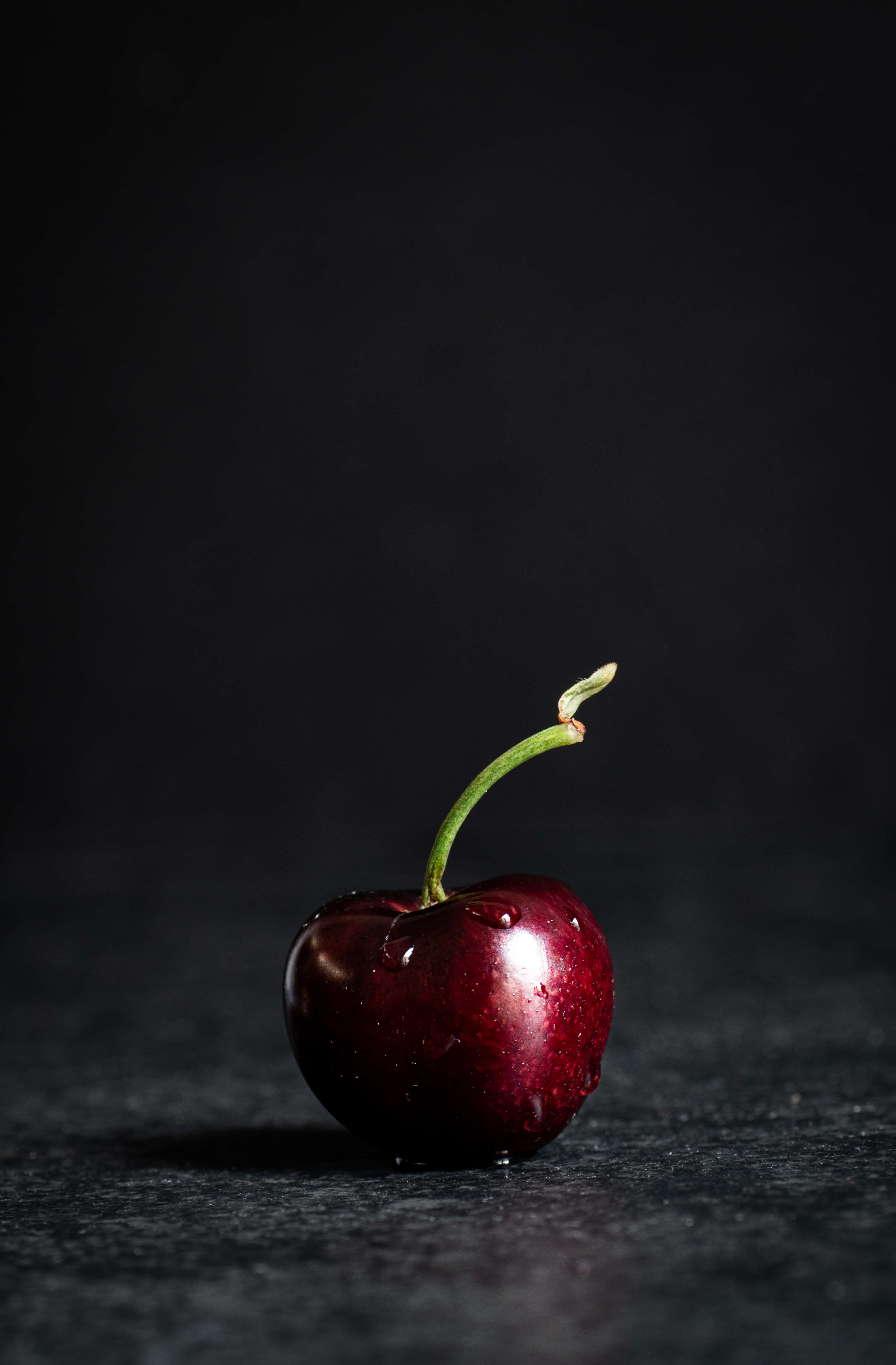Black Cherry 桌布 壁紙 — 一顆閃亮的紅櫻桃，帶有綠色莖，放在黑色背景的深色紋理表面上。