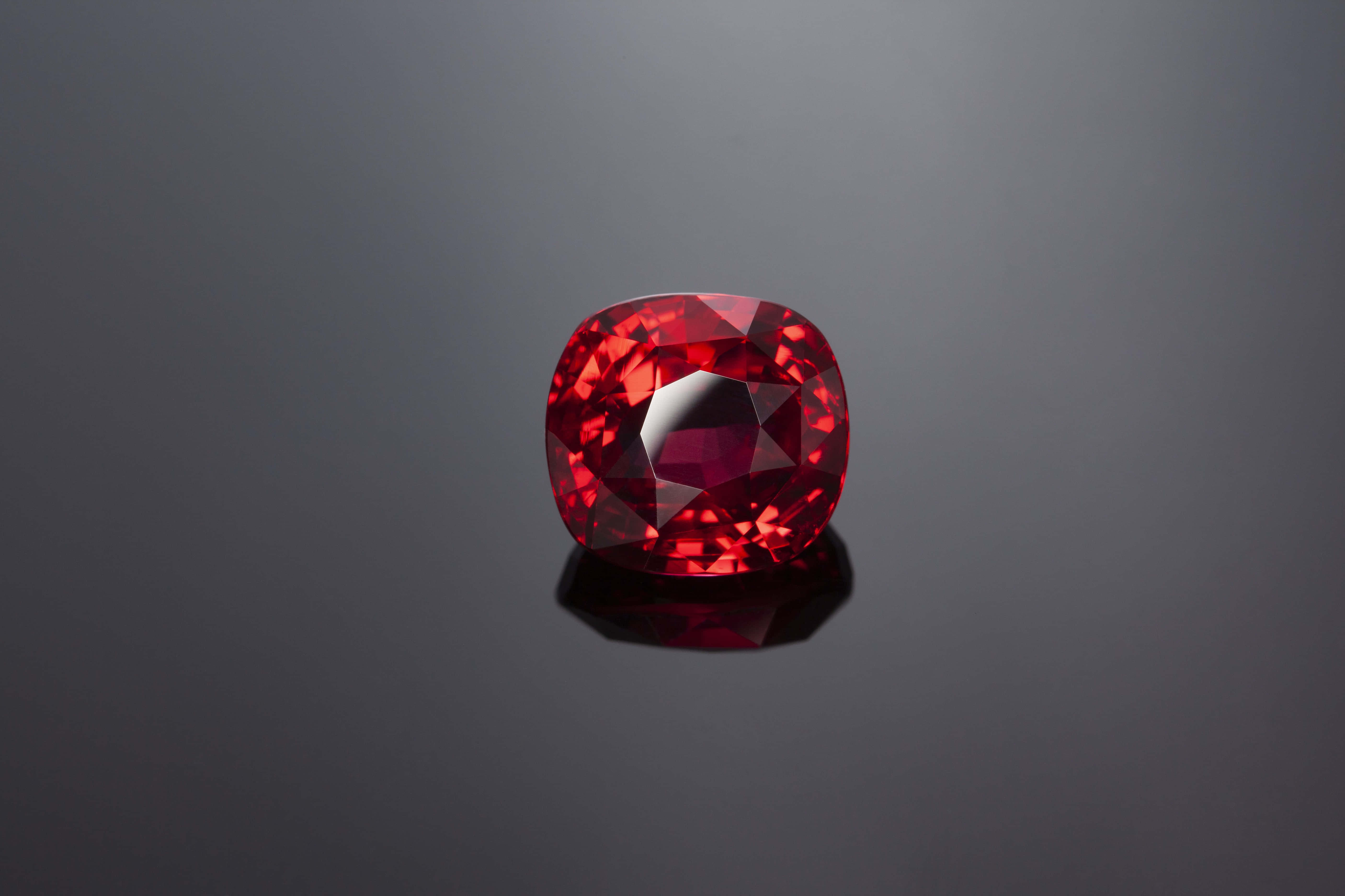 Ruby 桌布 壁紙 — 一顆閃耀的枕形切割紅寶石，在光滑的深灰色反光表面上閃爍著深紅色刻面。