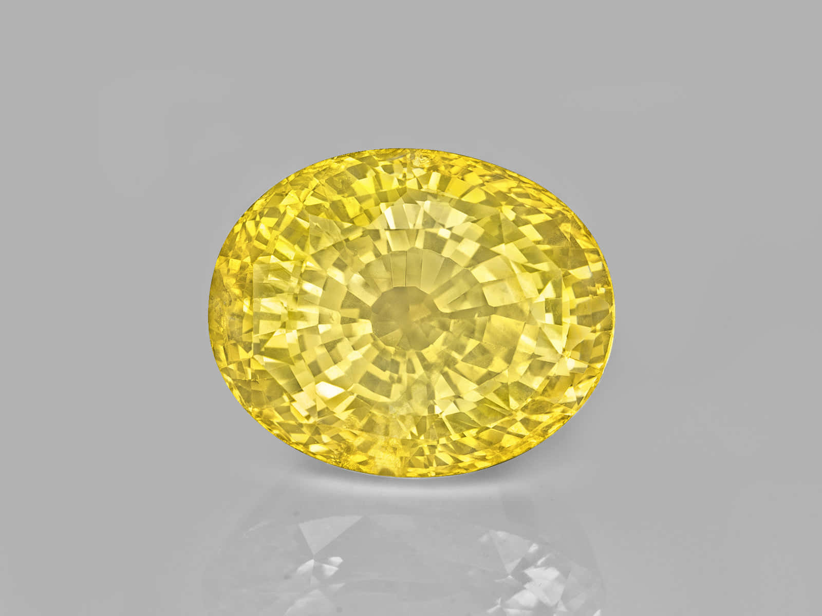 Yellow Sapphire 桌布 壁紙 — 一顆閃耀的黃色橢圓形切割寶石，帶有複雜的刻面，在灰色中性背景上閃閃發光。