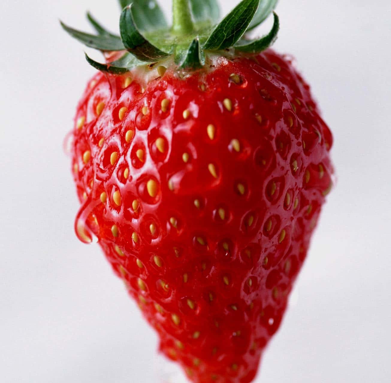 Red Strawberry 桌布 壁紙 — 一顆鮮紅色士多啤梨的特寫，帶有綠色葉子和細小的黃色種子，閃爍著水滴。