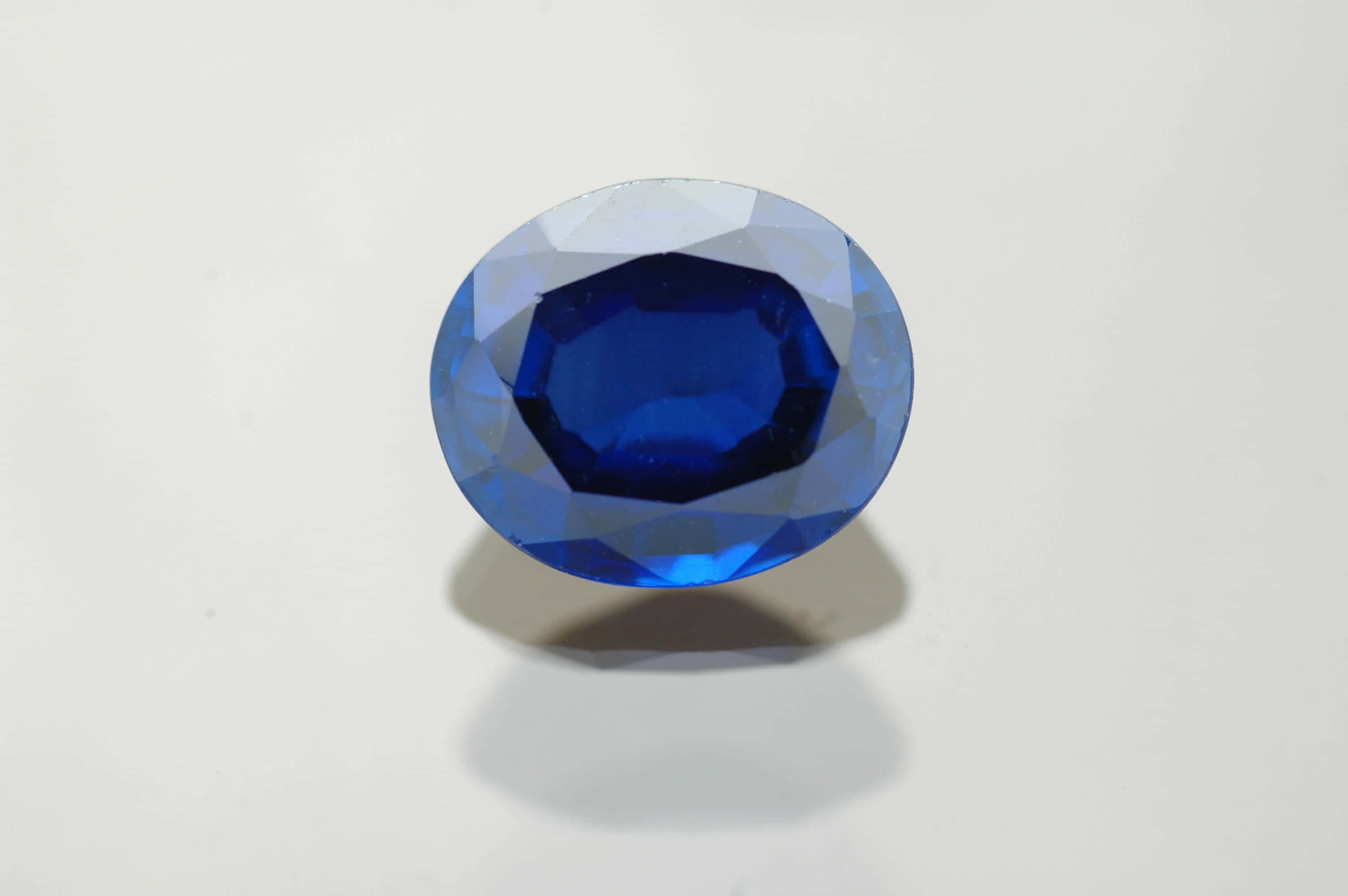 Sapphire Blue 桌布 壁紙 — 一顆鮮豔的藍色橢圓形切割藍寶石，在純白背景上閃耀著精確的刻面。
