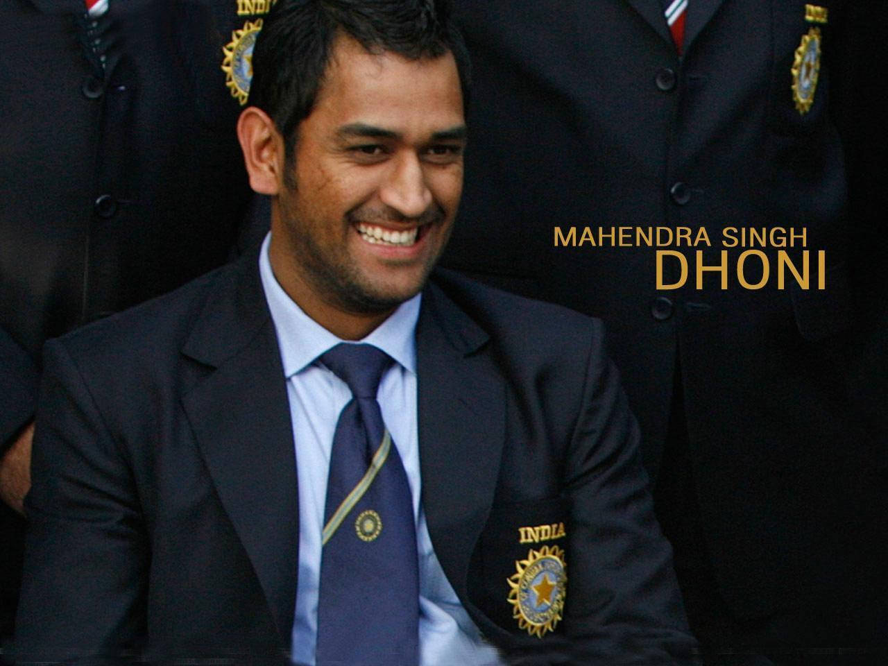 丁尼穿黑色西裝 壁紙 — Mahendra Singh Dhoni 身穿深色西裝，帶有印度板球隊徽章，背景是模糊的球隊背景，容光煥發。