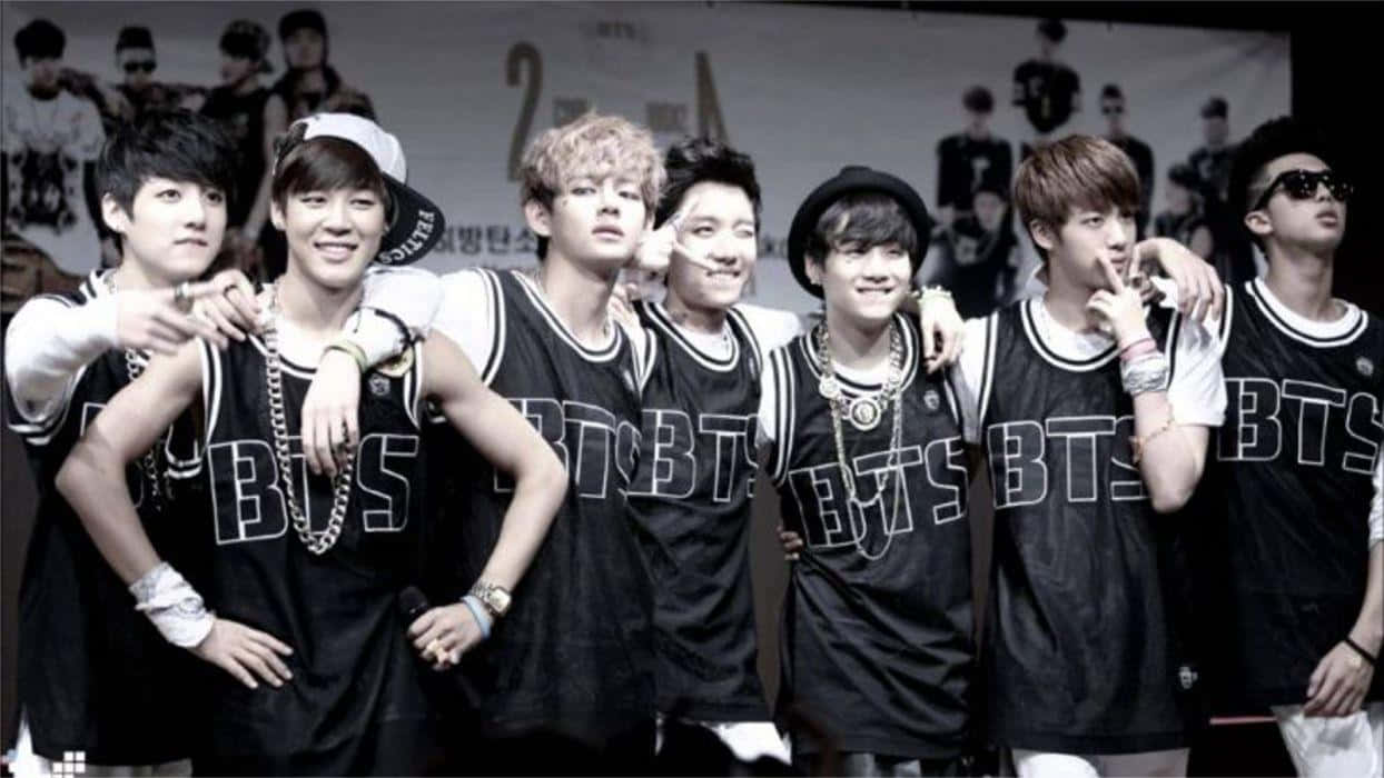 Bangtan Sonyeondan 桌布 壁紙 — 七位年輕男子身穿黑色 'BTS' 球衣，配戴飾物，表情各異，自信地擺出姿勢，背景為他們的標誌。