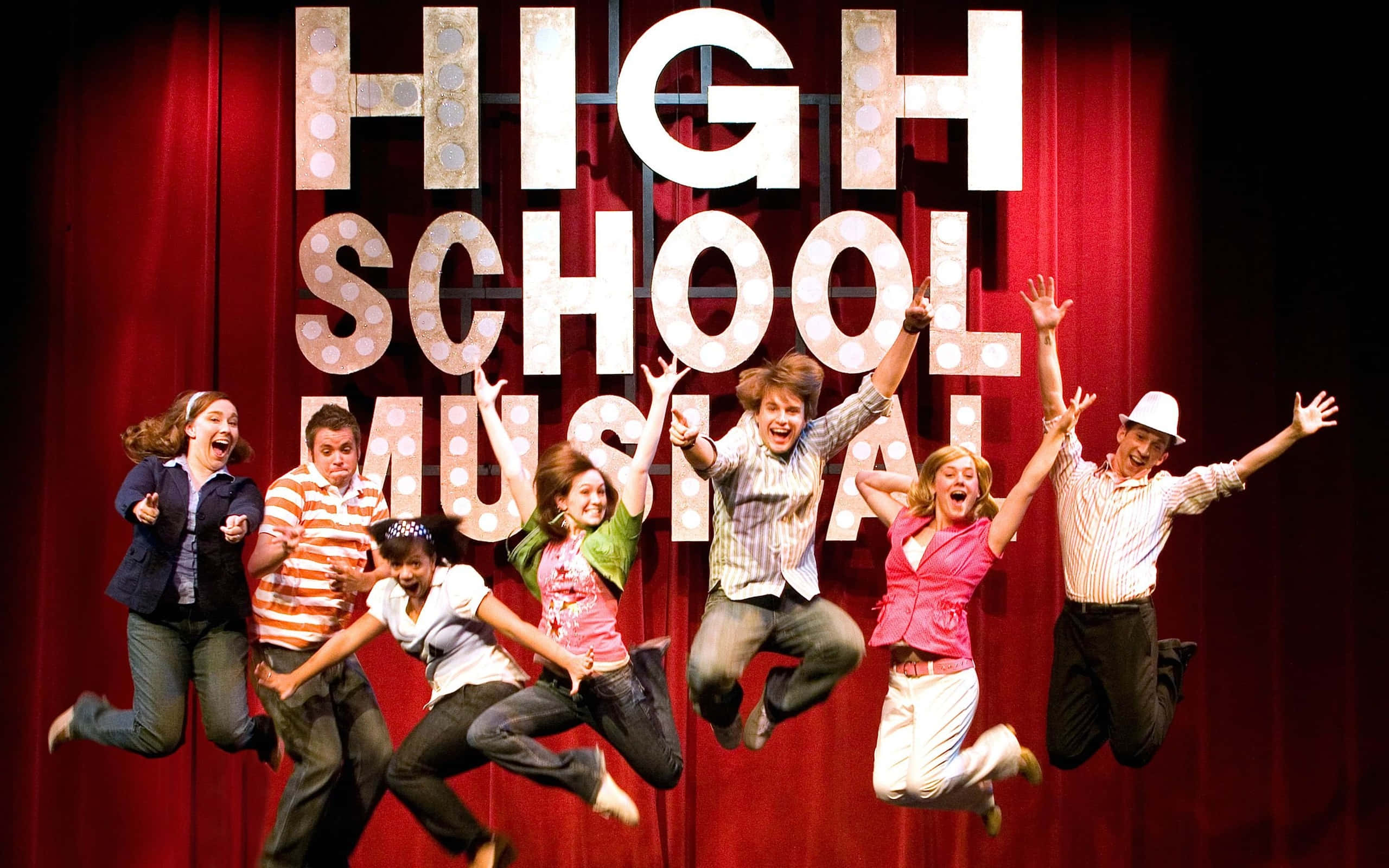 High School Musical 桌布 壁紙 — 七位快樂的演員在紅色帷幕前，於閃耀的「HIGH SCHOOL MUSICAL」標誌前，在舞台上雀躍地跳躍。