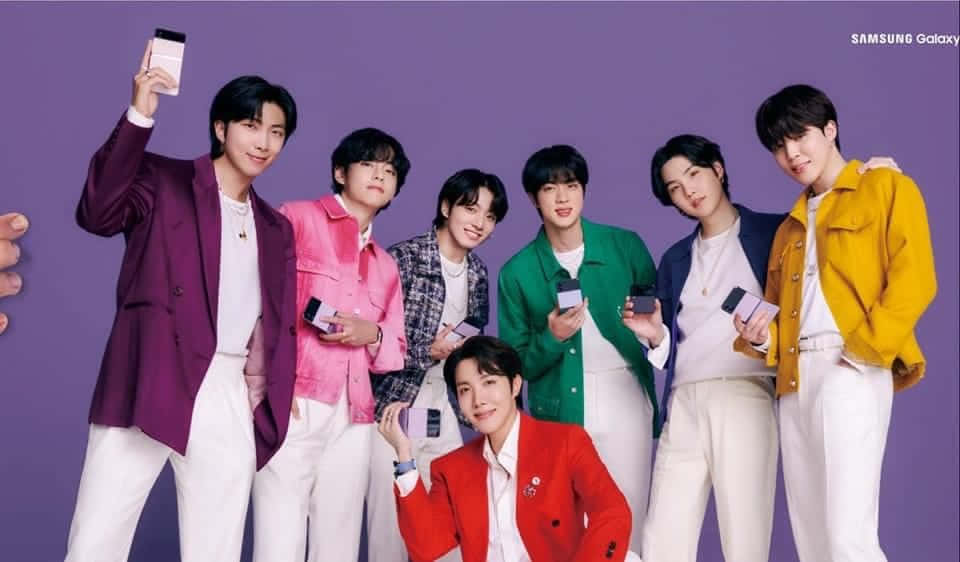 Bts Endorsements 桌布 壁紙 — 七位時尚年輕男子身穿彩色外套，在純紫色背景前愉快地擺姿勢，每人手持一部智能手機。