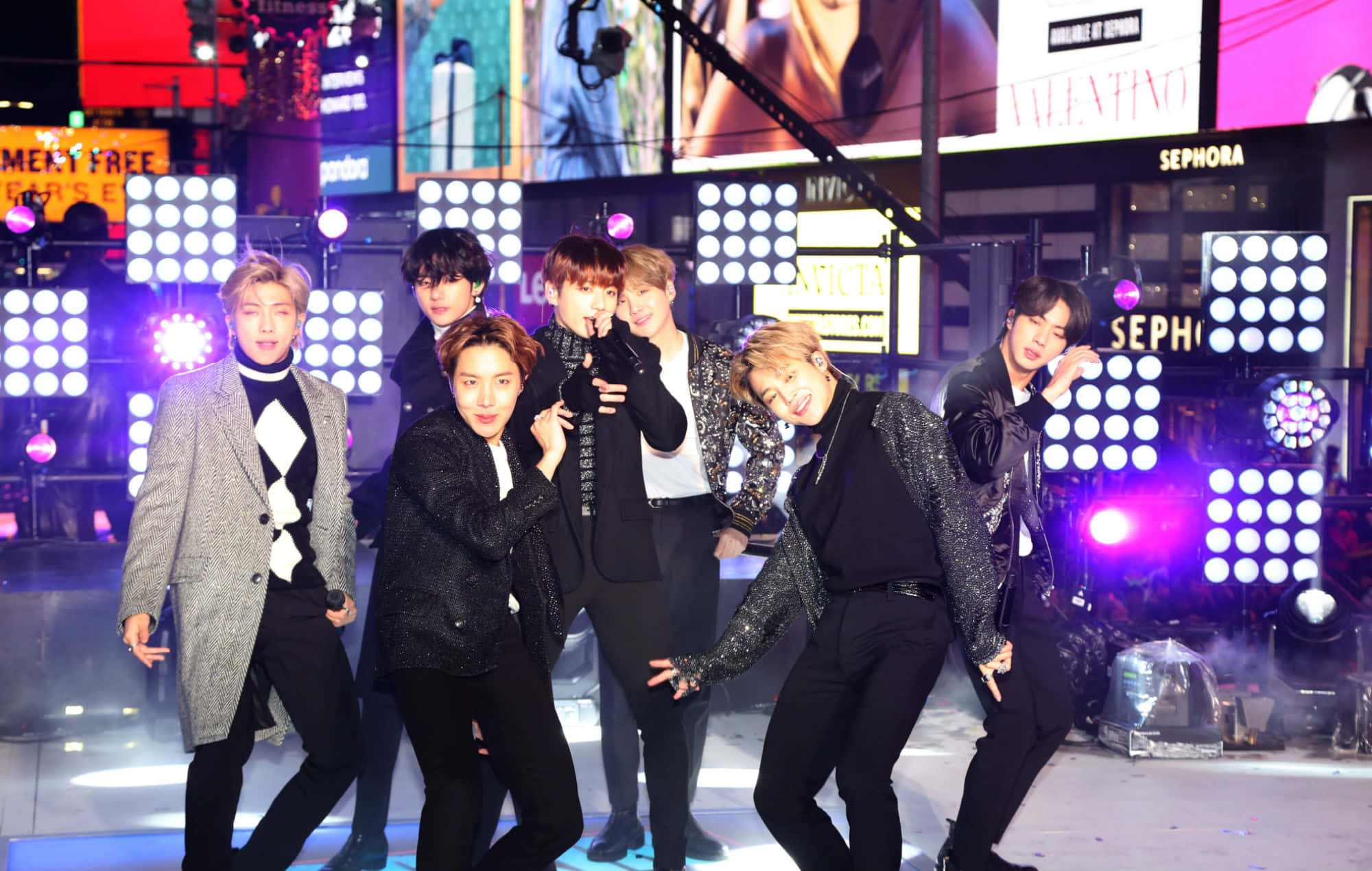 Bts Live 桌布 壁紙 — 七位時尚的男性表演者在明亮的舞台上擺出動態姿勢，身後有發光的方形燈光和城市廣告牌。