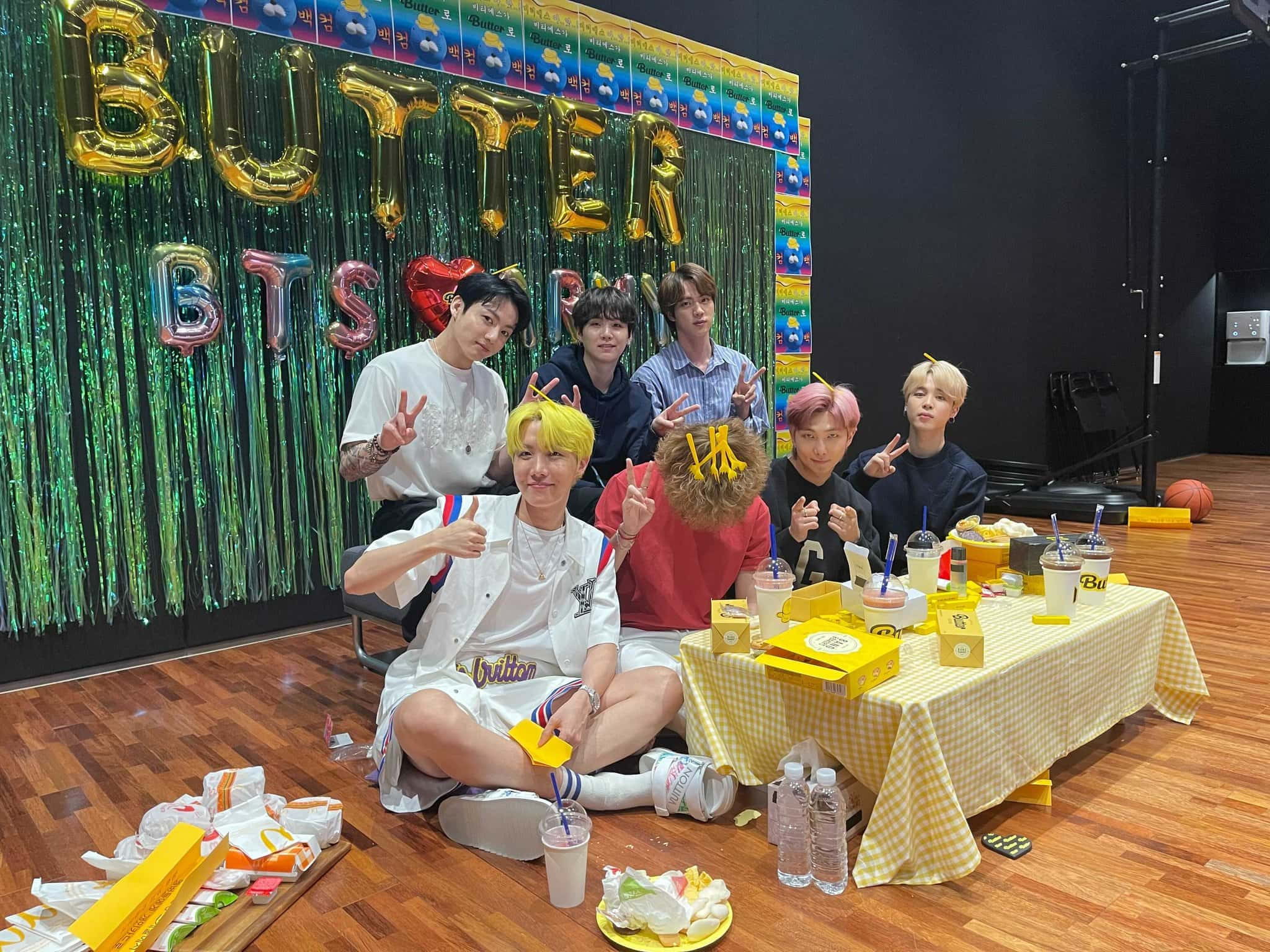Bts Butter 桌布 壁紙 — 七位身穿休閒服的年輕男子，圍著一張黃色格仔枱，手持飲品和小食，在閃爍的綠色背景前，與「BUTTER BTS」氣球愉快地擺姿勢。