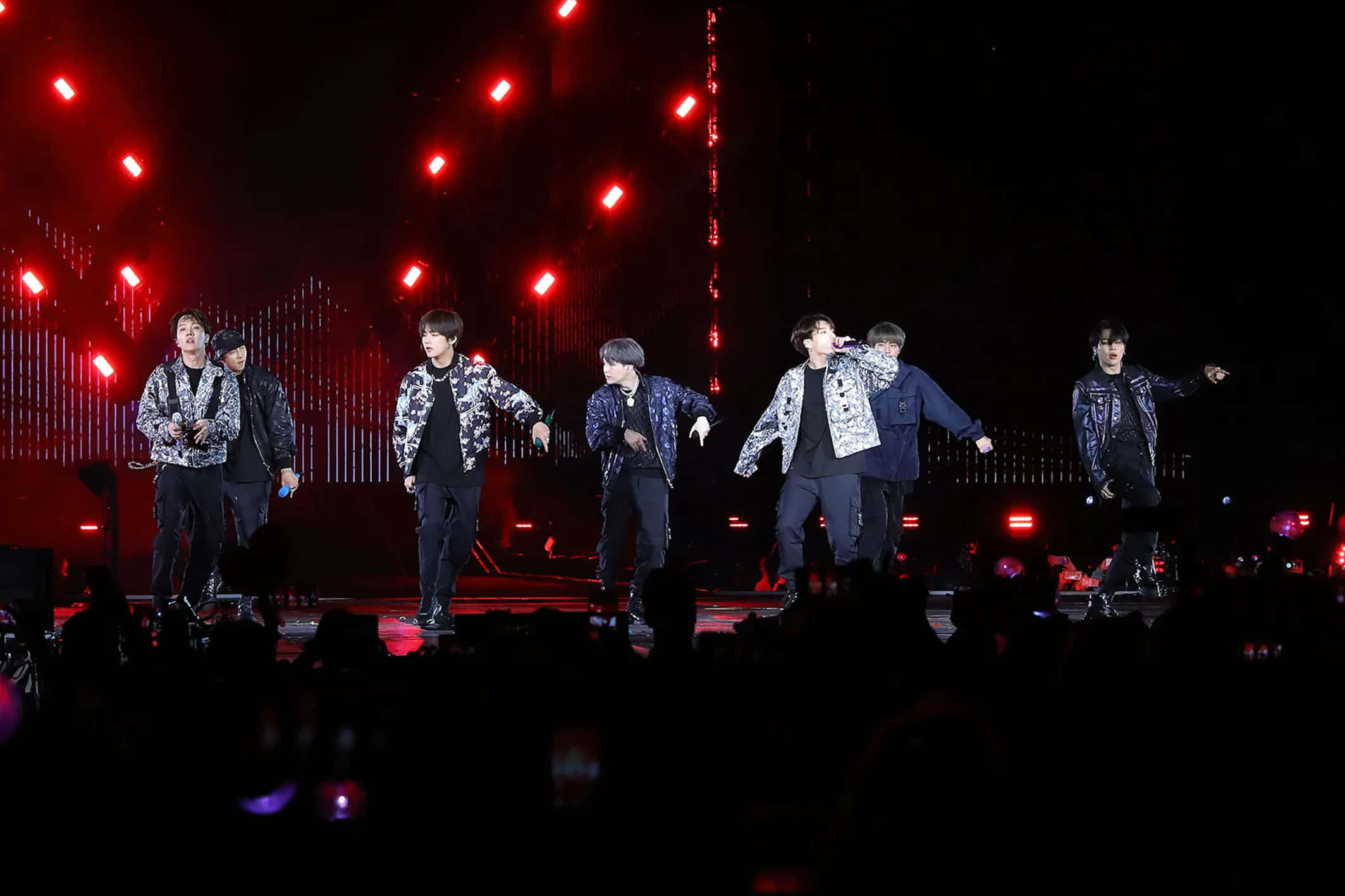 Bts Stage 桌布 壁紙 — 七位身穿圖案外套的男表演者在紅色舞台燈光下充滿活力地跳舞，前景有黑暗的觀眾。
