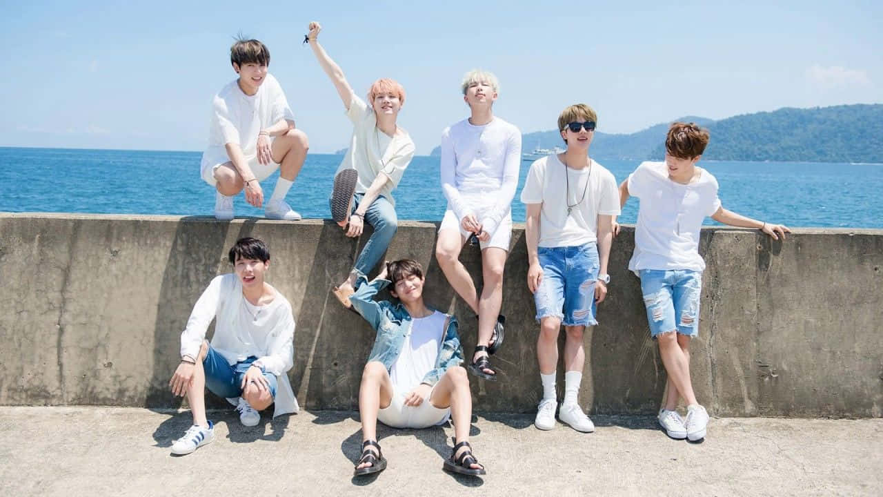 Bangtan Sonyeondan 桌布 壁紙 — 七位身穿白襯衫和牛仔短褲的年輕男子，在明亮的藍色海洋和天空背景下，隨意地擺姿勢在混凝土海堤上。