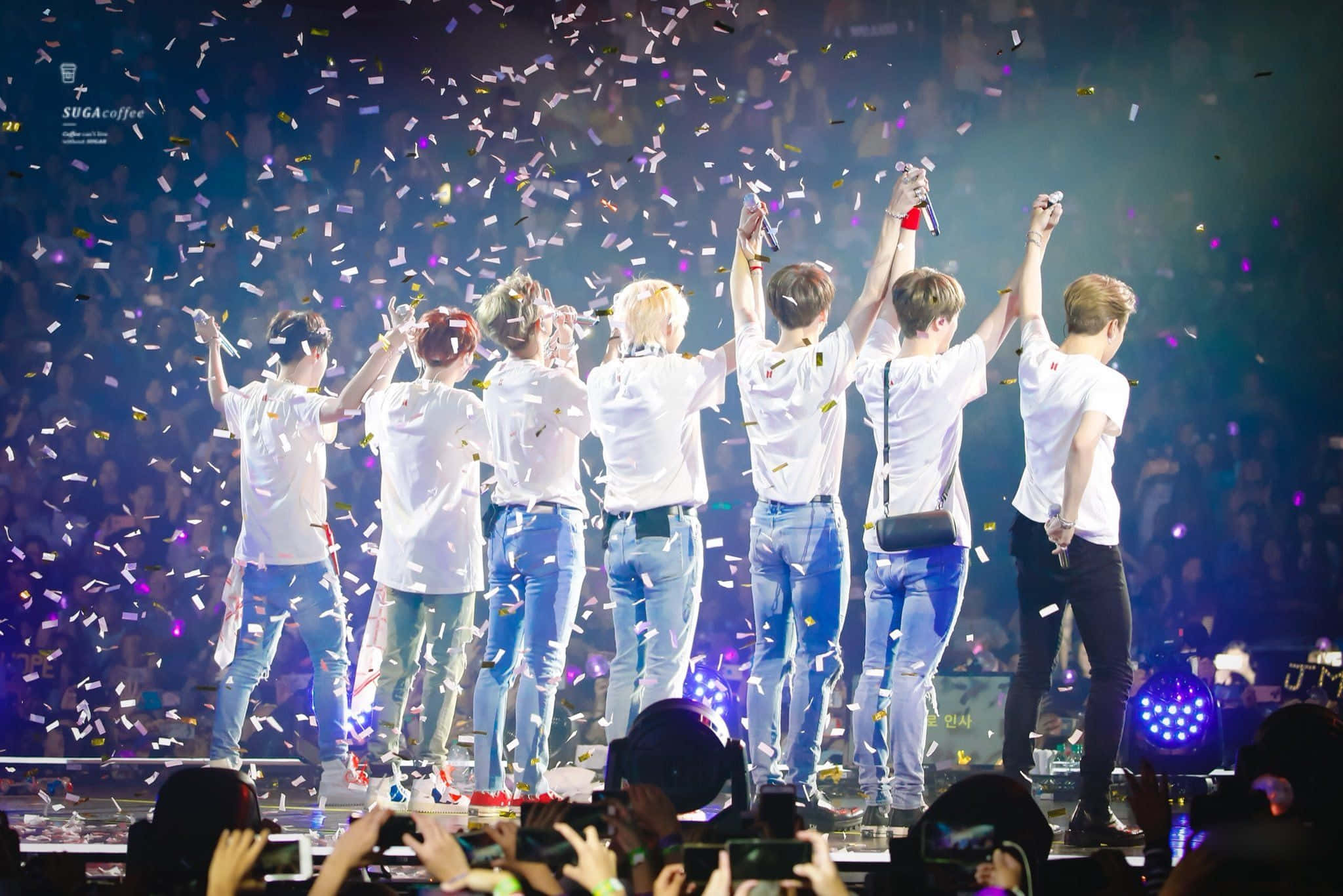 Bts Concert 桌布 壁紙 — 七位身穿白襯衫和牛仔褲的表演者背光站在舞台上舉起雙臂，彩色紙屑如雨般灑落在歡呼的粉絲身上。
