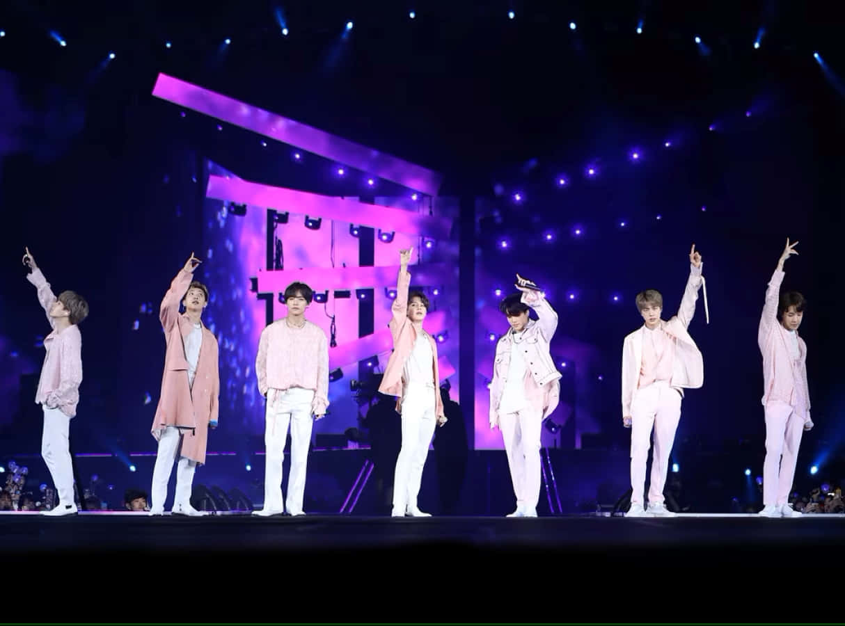 Bts Stage 桌布 壁紙 — 七位身穿粉紅和白色服裝的男表演者在充滿活力的紫色舞台燈光下向粉絲揮手。