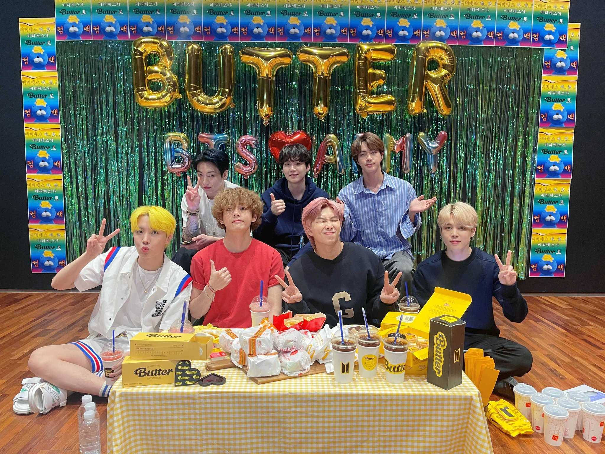 Butter 桌布 壁紙 — 七位身穿色彩繽紛服裝的年輕男性，在擺滿牛油主題小食和飲品的桌子後，對著閃爍的綠色背景愉快地擺姿勢。