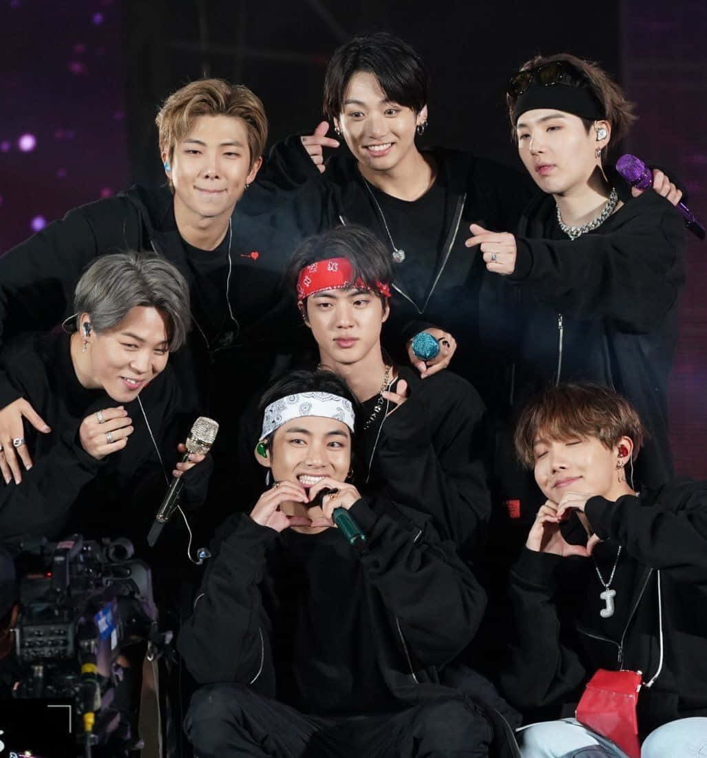 Bts Concert 桌布 壁紙 — 七位身穿黑色服裝、面帶微笑的K-pop偶像在紫色燈光和麥克風的舞台上一起擺姿勢。