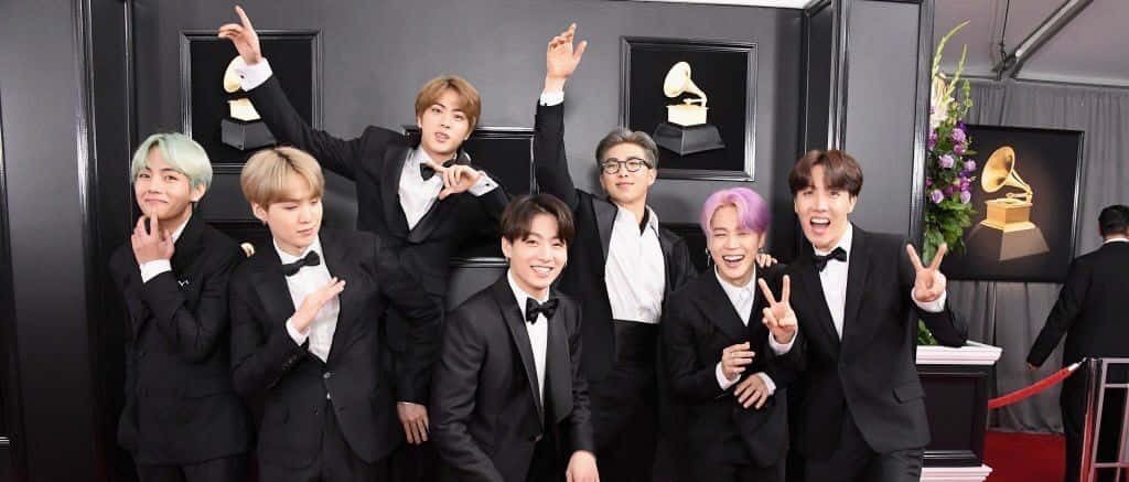 Big Hit Entertainment 桌布 壁紙 — 七位身穿黑色燕尾服、頭髮色彩繽紛的男士在紅地毯上與格林美獎盃旁愉快地擺姿勢。