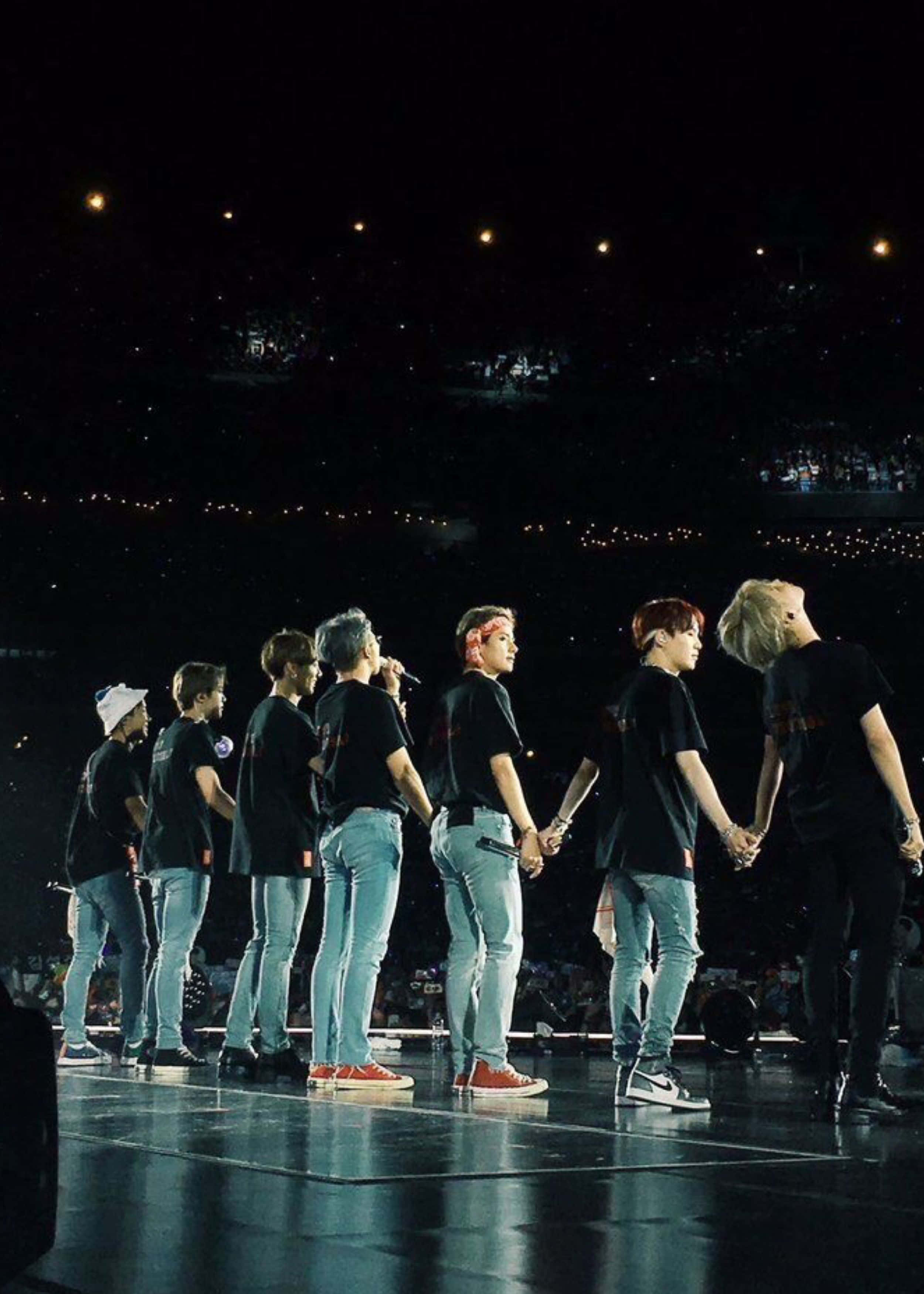 Bts Stage 桌布 壁紙 — 七位身穿黑色襯衫和牛仔褲的男性表演者在夜間音樂會中，在燈光璀璨的舞台上排成一排，手牽手，背景是黑暗的觀眾。