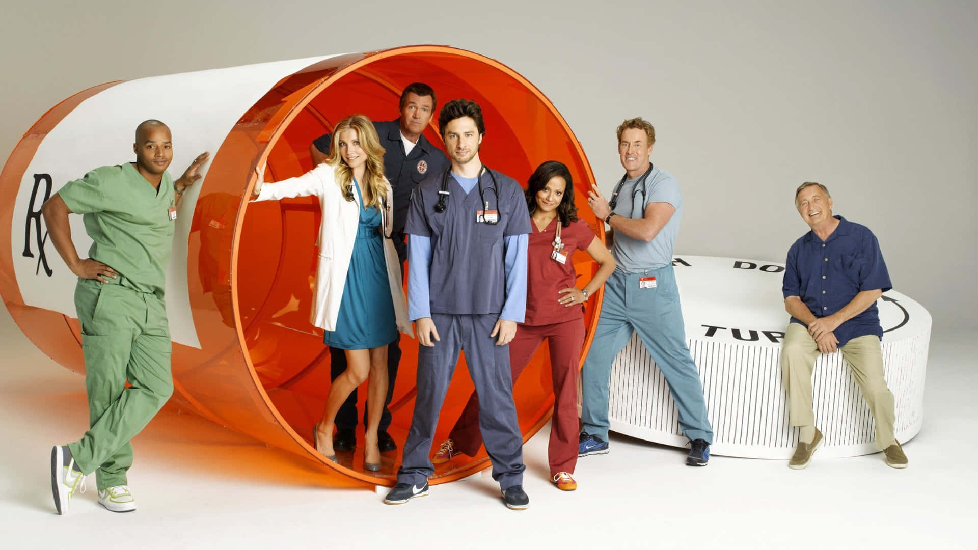 John C. Mcginley 桌布 壁紙 — 七位醫療電視劇演員在攝影棚內自信地圍繞一個巨大的橙色和白色藥丸瓶道具擺姿勢。