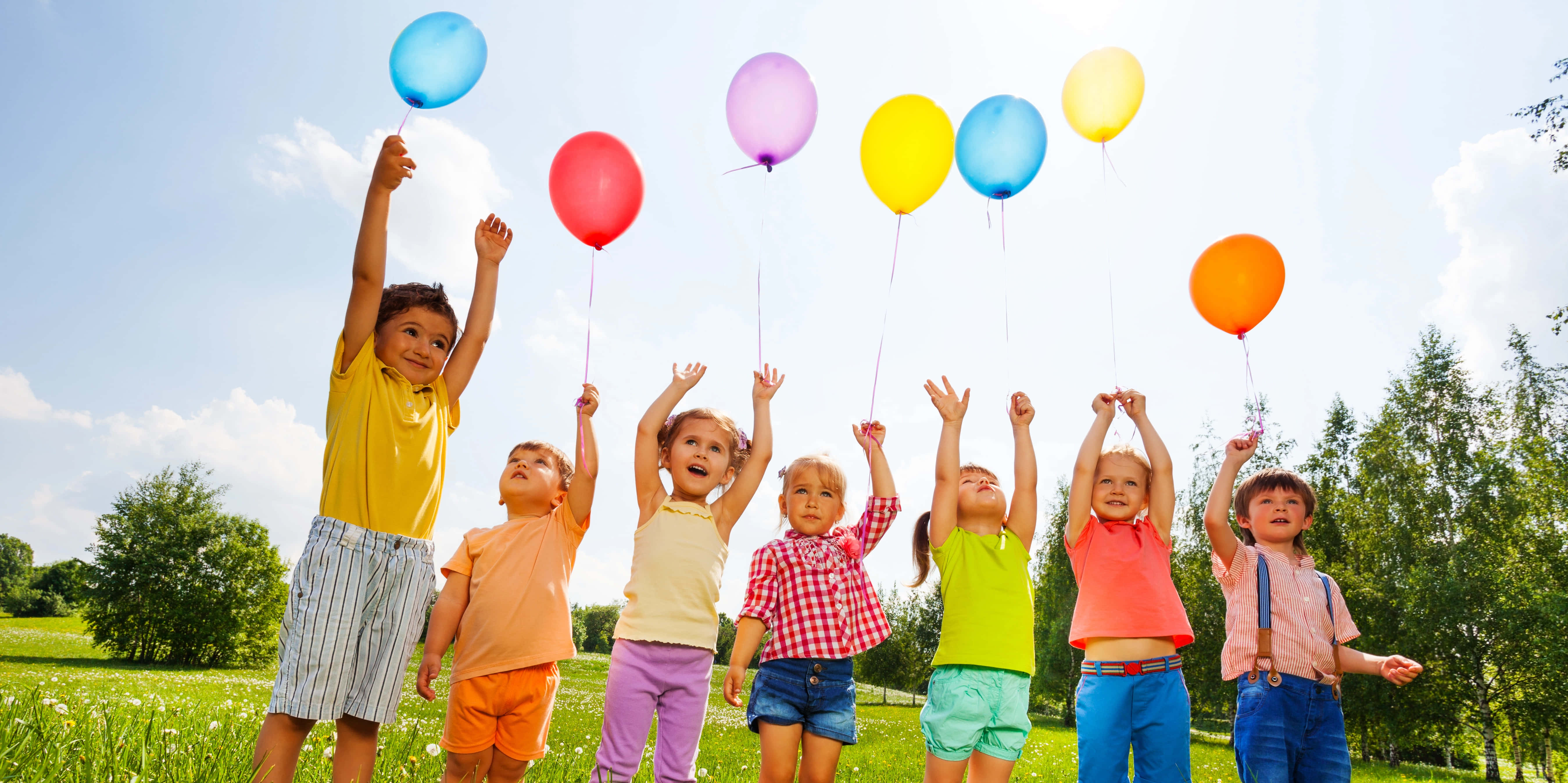 children 桌布 壁紙 — 七個快樂的孩子在陽光明媚的草地上，將色彩繽紛的氣球舉向明亮的藍天。