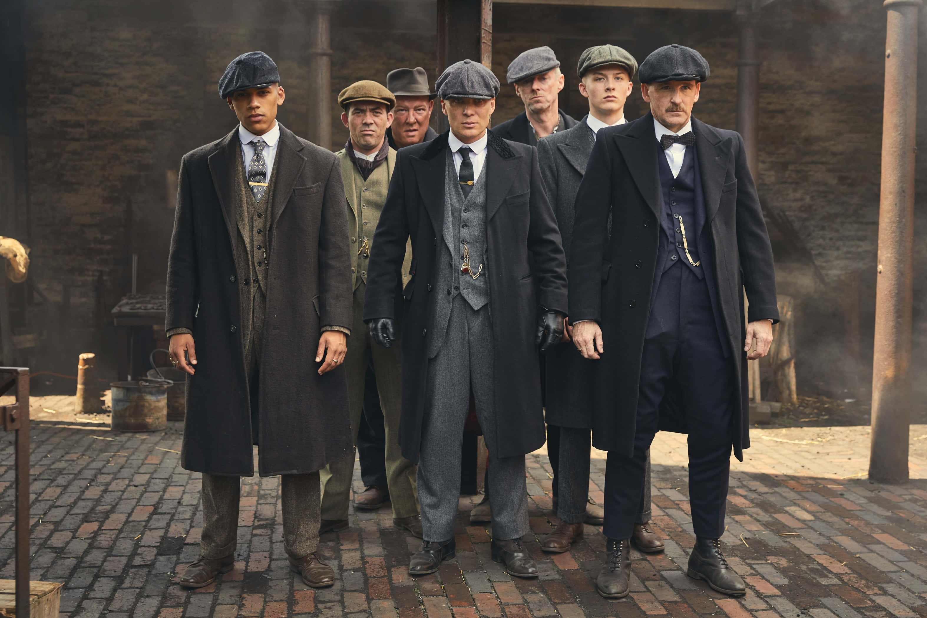 Peaky Blinders 桌布 壁紙 — 七個戴著復古平頂帽、身穿訂製西裝的男人，在磚砌庭院中莊嚴地站立，背景有煙霧飄散。